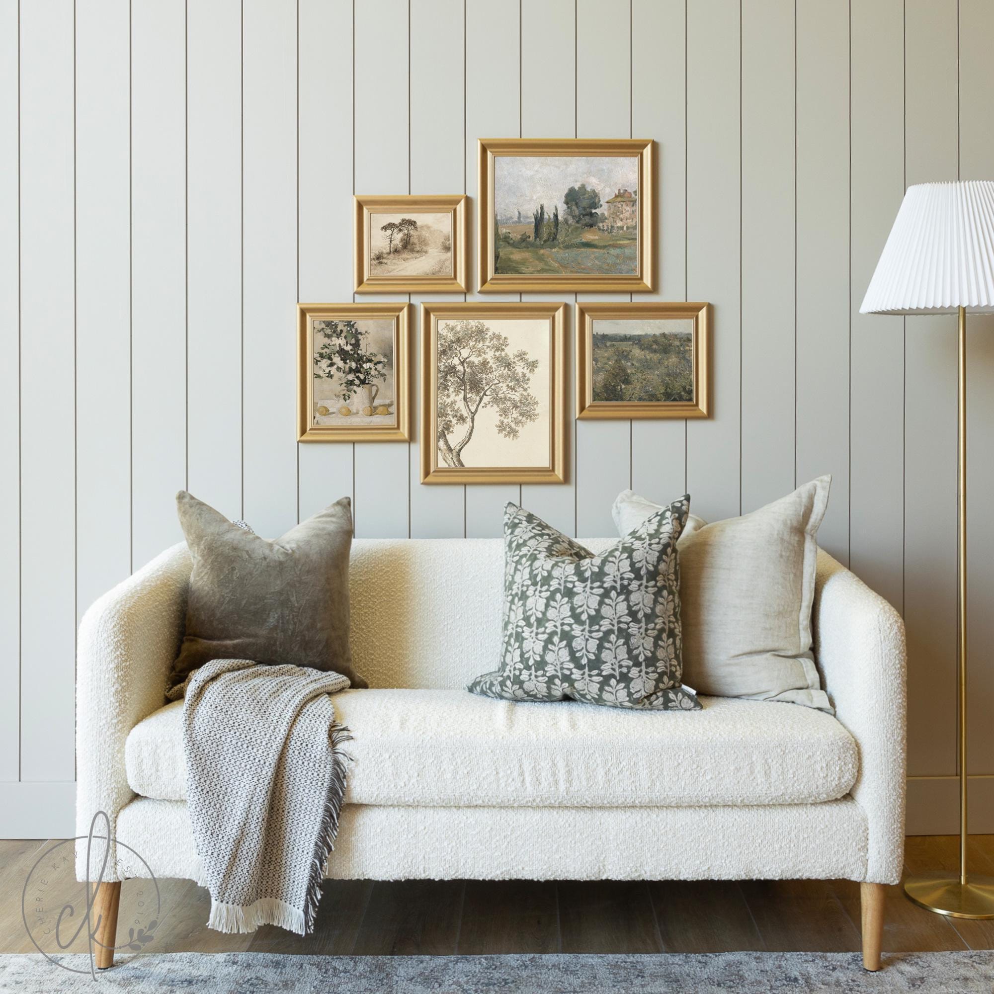Cozy living room featuring a white textured sofa with decorative pillows and a throw blanket, accented by a gold-framed gallery wall set of vintage-inspired artwork, including landscapes, botanicals, and still-life pieces, against a shiplap wall.