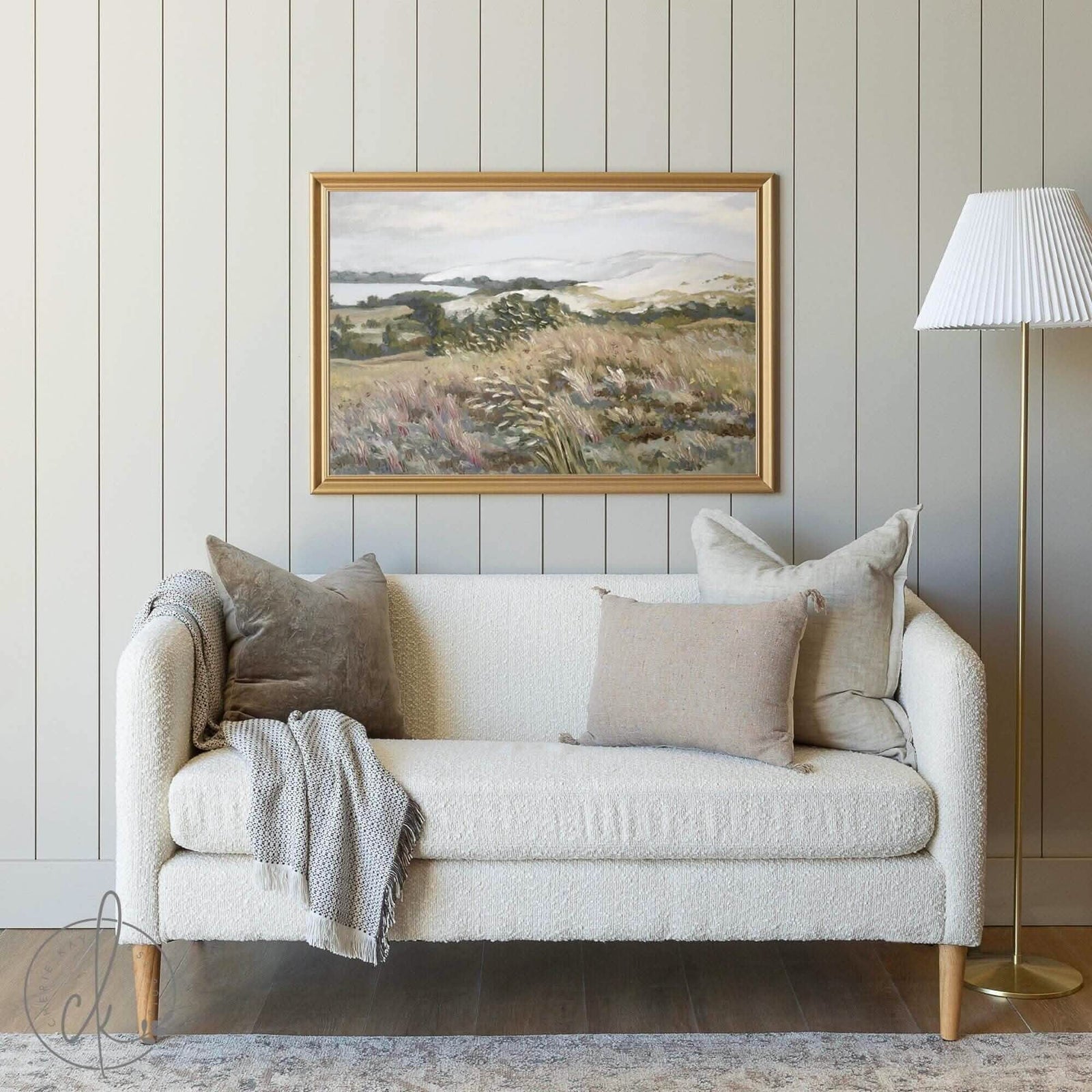 A cozy living room with a beige textured couch, neutral pillows, and a gray knit throw. A gold-framed landscape painting hangs on a light gray paneled wall. A white-shaded floor lamp stands to the right, creating a serene and minimalist space.