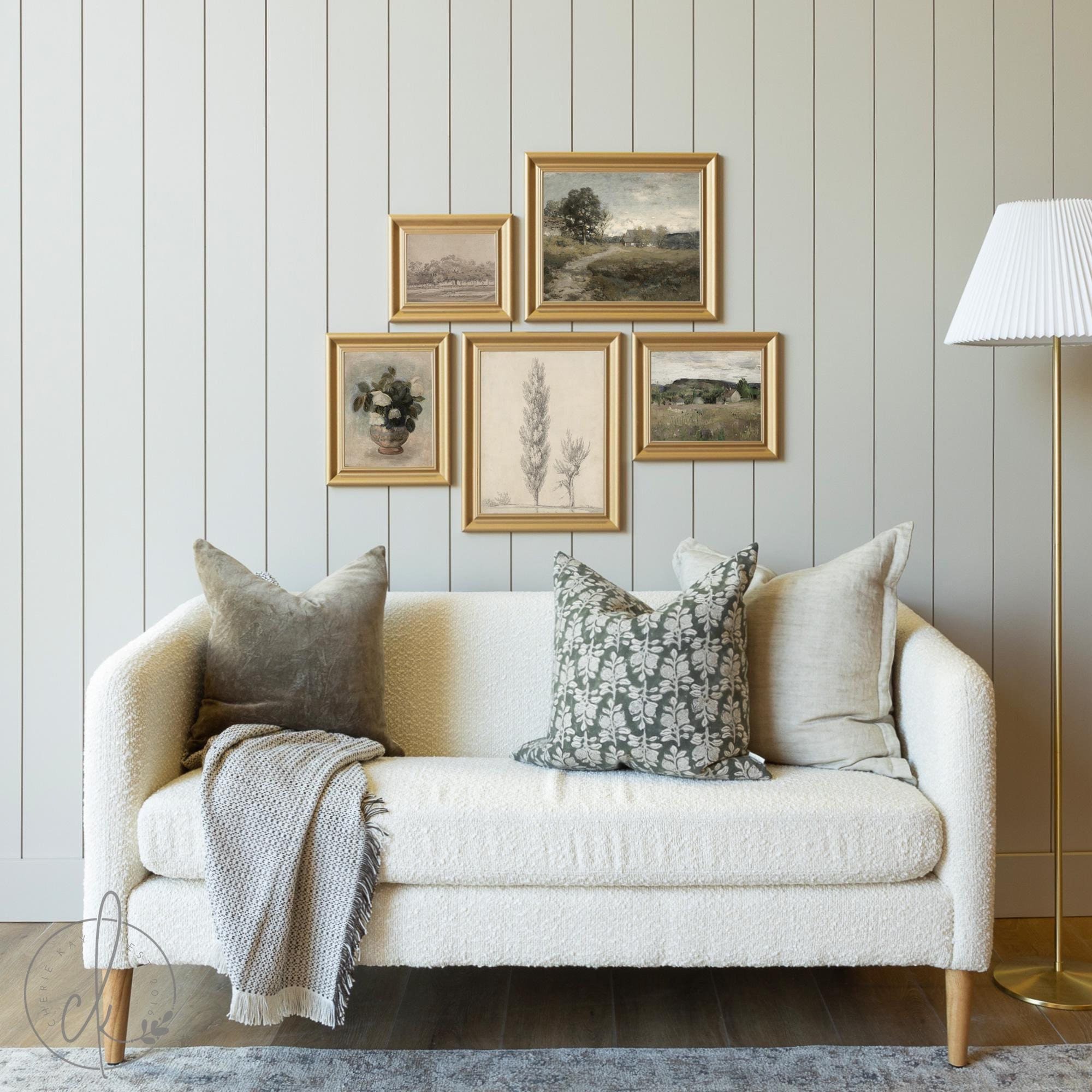 A cozy living room setup featuring a modern farmhouse-style gallery wall with five framed nature-inspired art prints, a cream textured sofa adorned with neutral and floral-patterned pillows, a soft throw blanket, and a standing lamp in the corner.