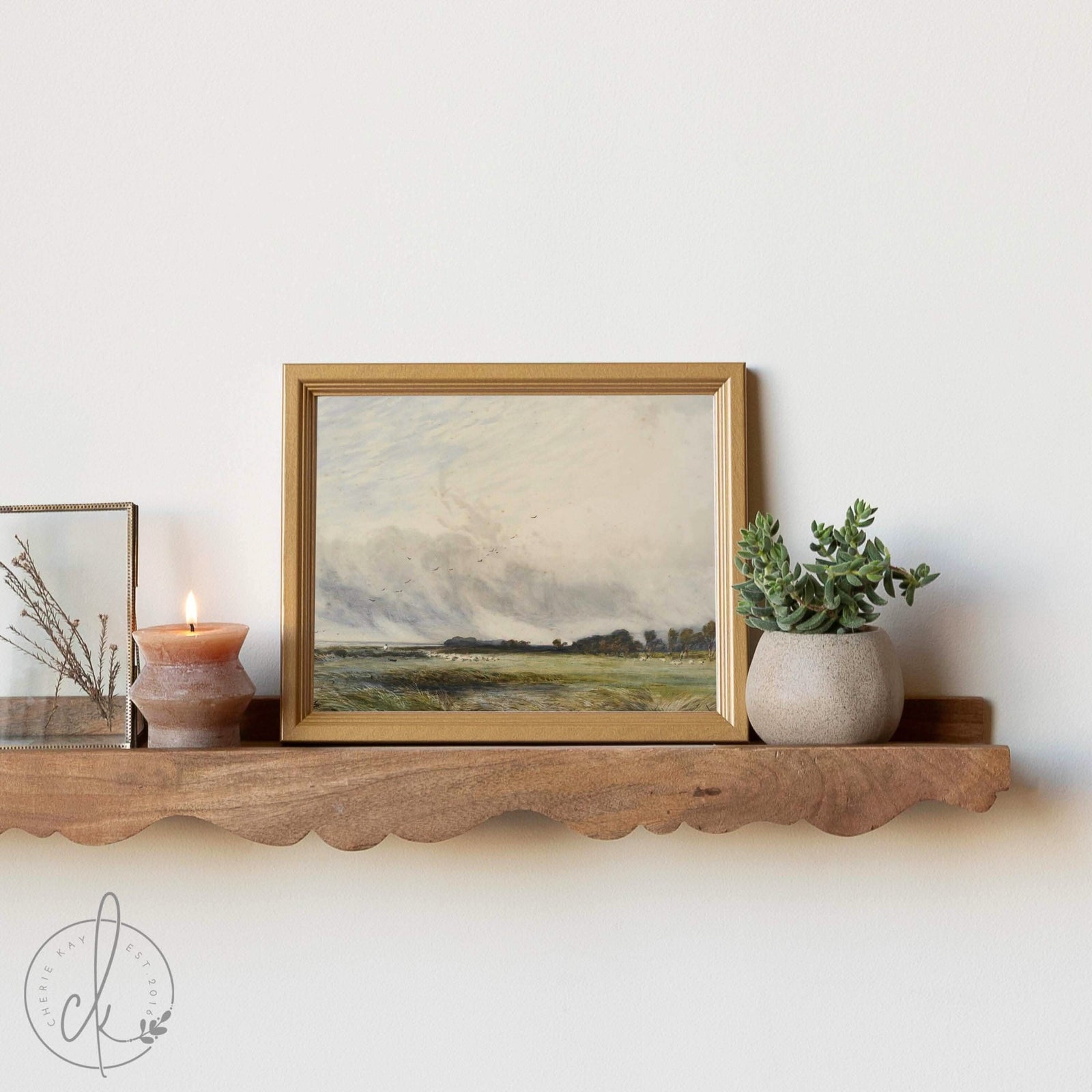 A small framed landscape painting on a wooden floating shelf, accompanied by a lit candle, a potted succulent, and a glass frame with dried botanicals, creating a cozy, minimalist decor arrangement.