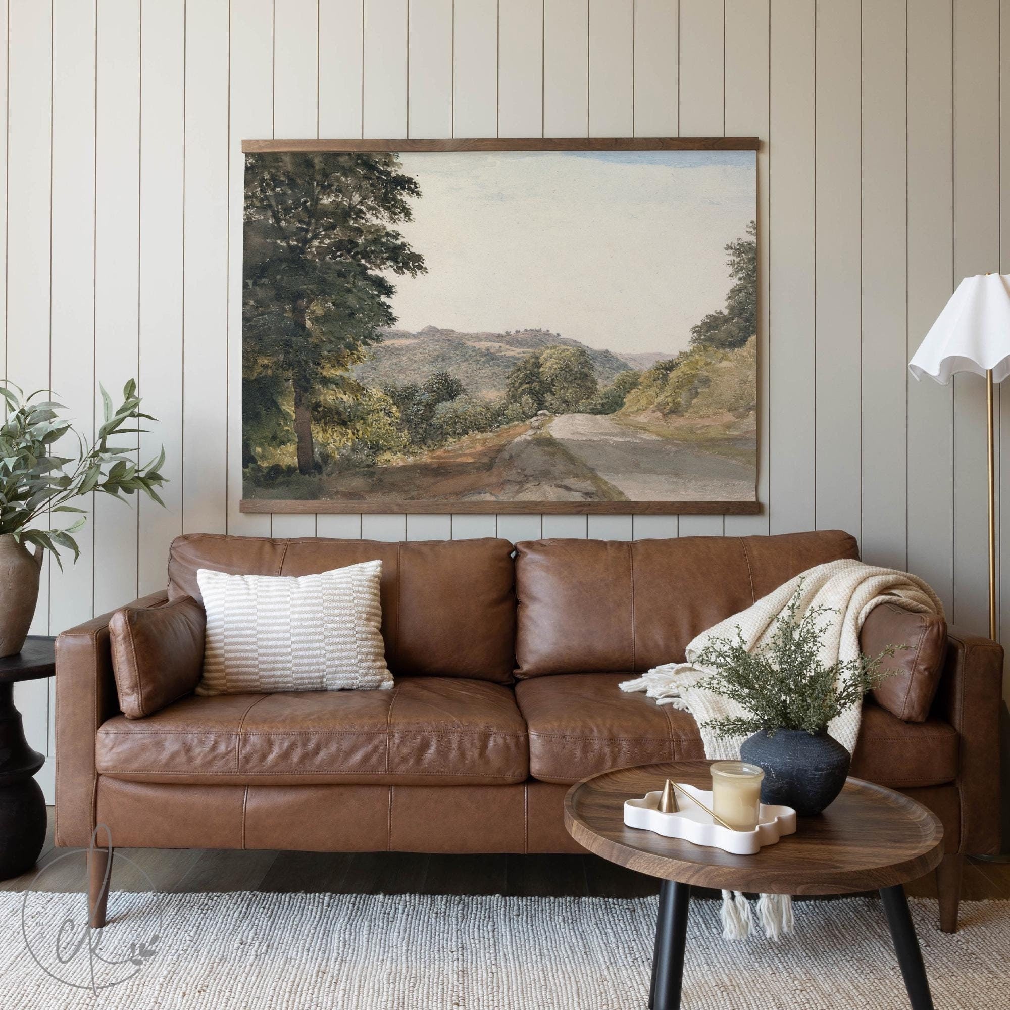 A cozy living room with a brown leather sofa, a textured pillow, and a blanket draped over the arm. A round wooden coffee table with simple decor sits in front, and a wall-mounted art piece of a scenic road and trees hangs above.