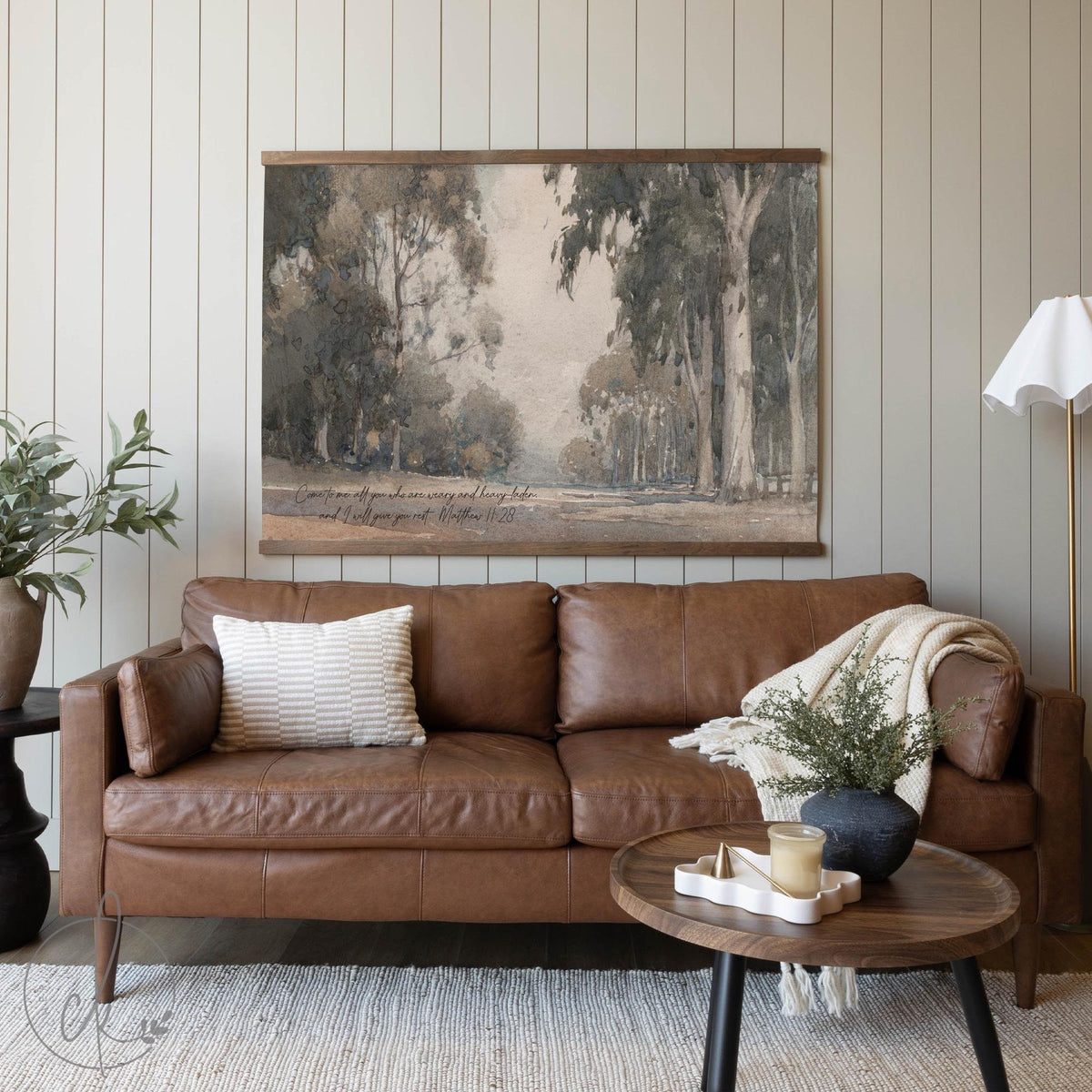 A cozy living room featuring a brown leather sofa, a textured pillow, and a draped blanket. A wooden coffee table with decor sits in front, and an art piece of a tranquil tree-lined path with a scripture verse from Matthew 11:28 hangs above.