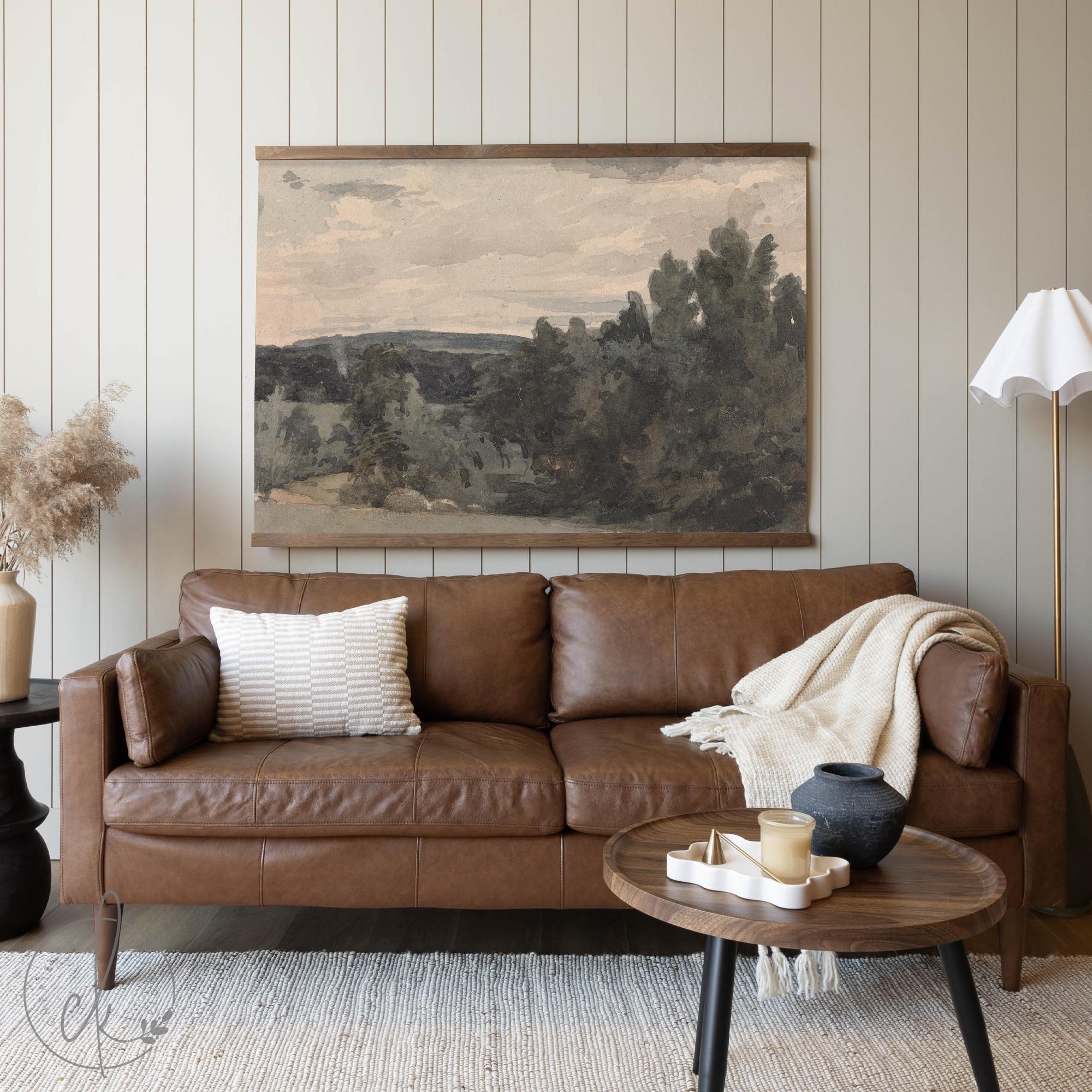 &quot;A warm, neutral-toned living room featuring a brown leather sofa, a large landscape tapestry with soft muted colors hanging above, and simple decor accents including a cream throw, decorative pillows, and a small round wooden coffee table.&quot;