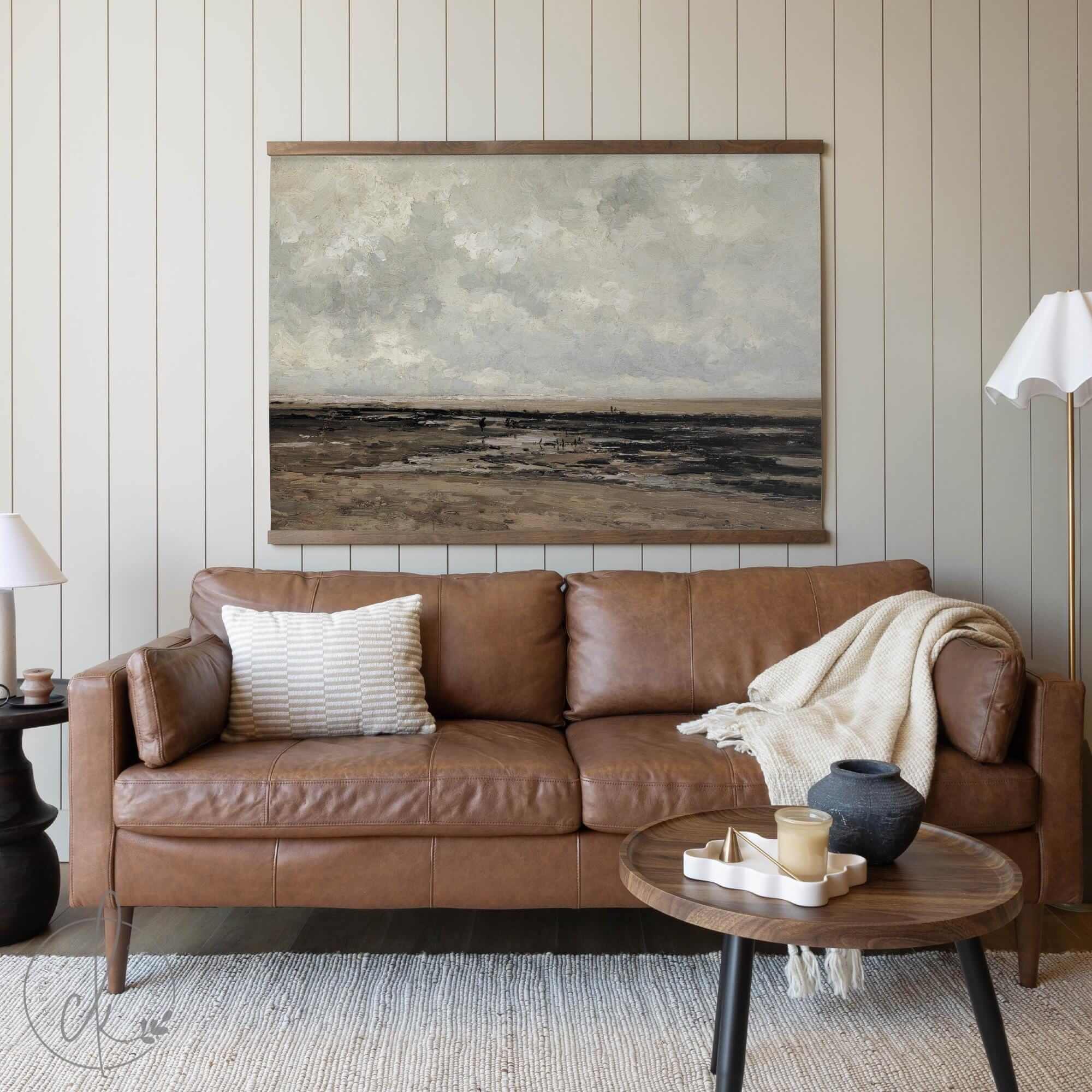 Neutral-toned landscape tapestry wall art featuring a vintage coastal scene with textured brushstrokes, framed with a wooden hanger, displayed above a brown leather sofa in a cozy living room setting.