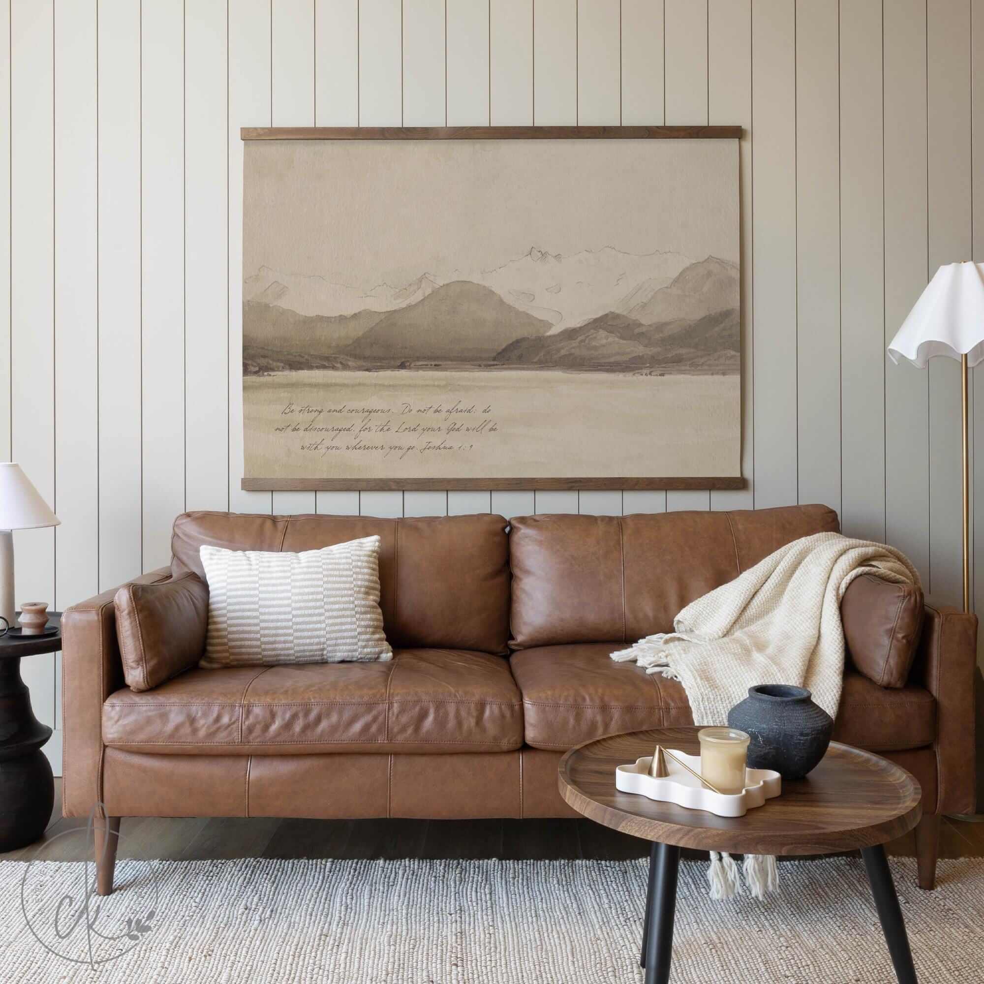Large vintage-style landscape tapestry featuring muted mountain scenery and an inspirational Bible verse quote, hanging above a brown leather couch in a neutral-toned living room with cozy decor.