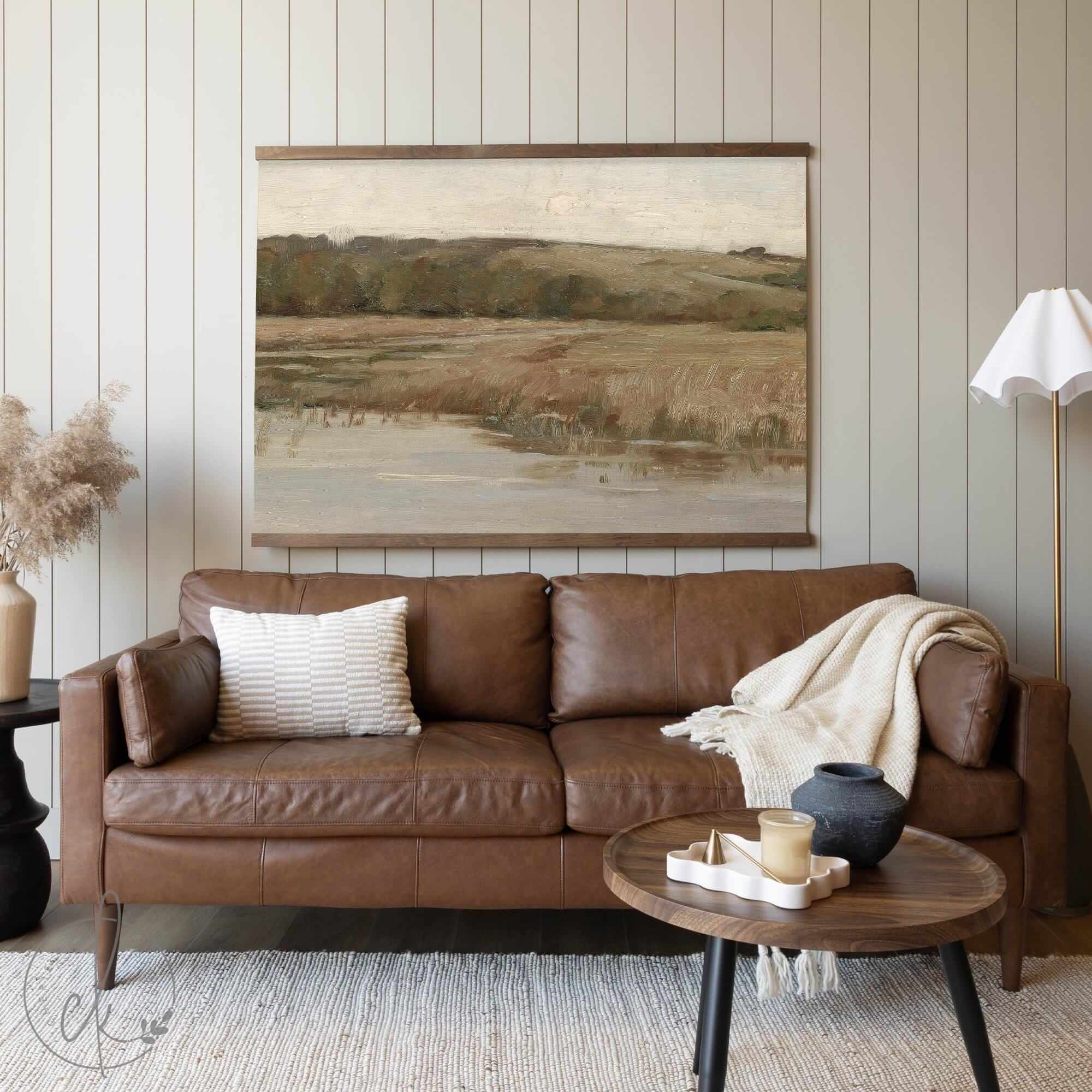A large, neutral-toned landscape tapestry featuring rolling hills and grassy fields, hung above a warm brown leather sofa in a cozy living room setting.