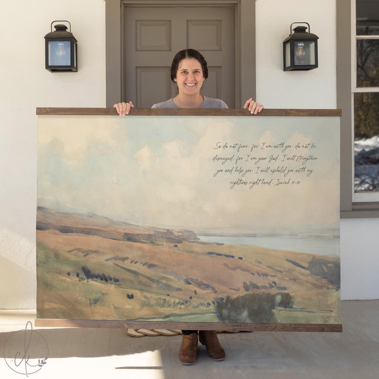 Woman holding a large tapestry featuring a watercolor landscape and the Bible verse Isaiah 41:10, displayed in front of a house door.