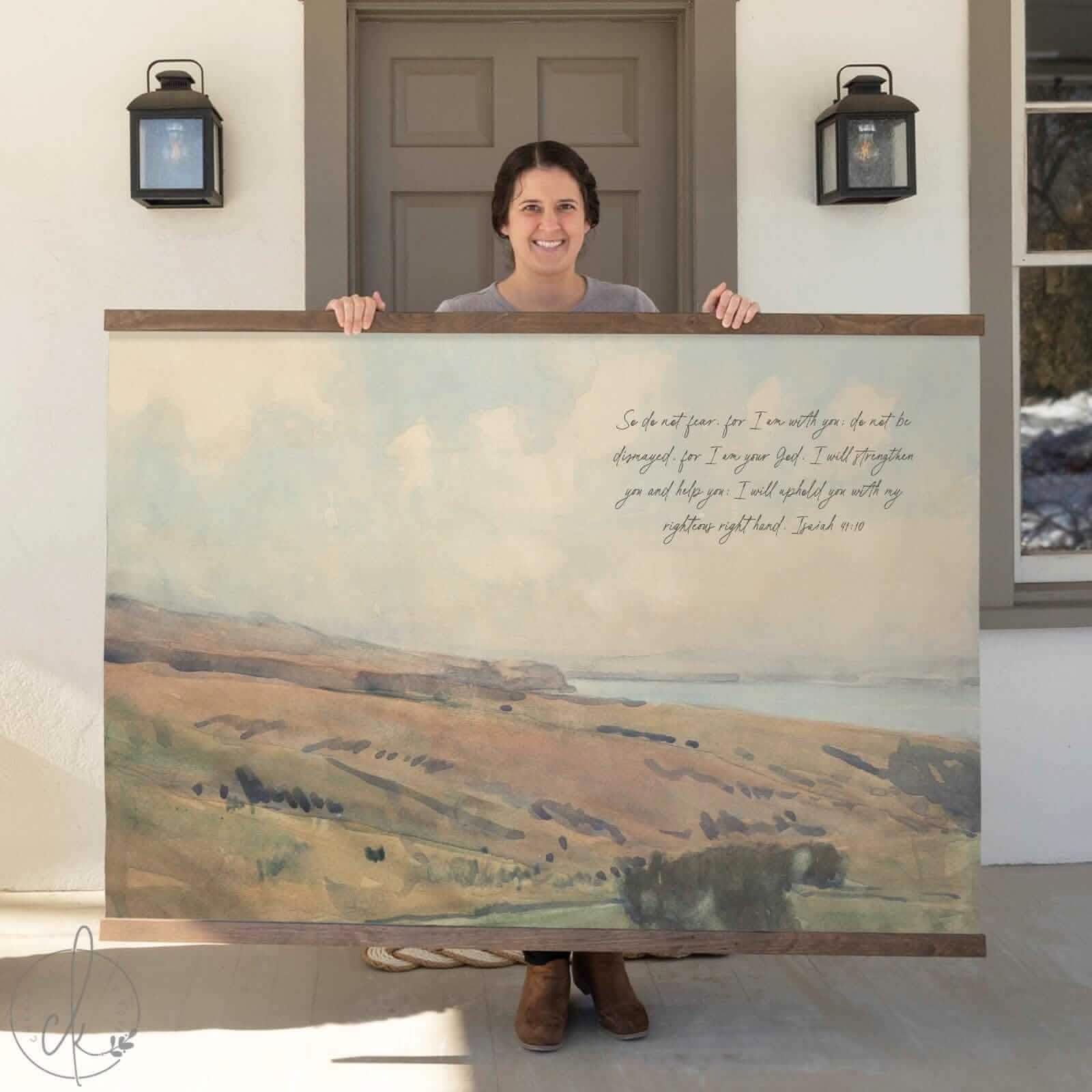 Woman holding a large tapestry featuring a watercolor landscape and the Bible verse Isaiah 41:10, displayed in front of a house door.