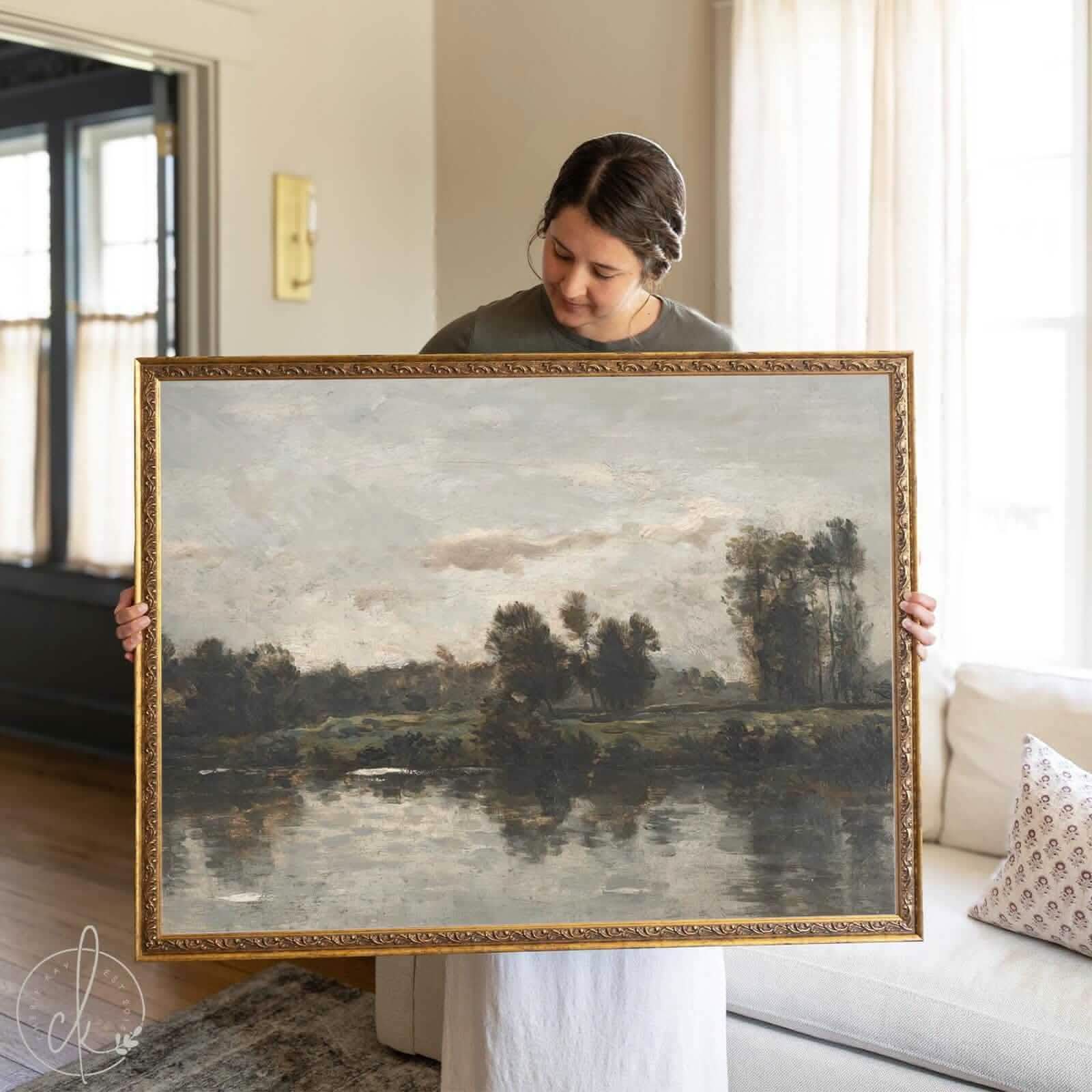 Woman holding a framed vintage oil painting of a moody river landscape with trees, cloudy sky, and ornate gold frame—ideal vintage wall art for traditional or modern vintage home decor.