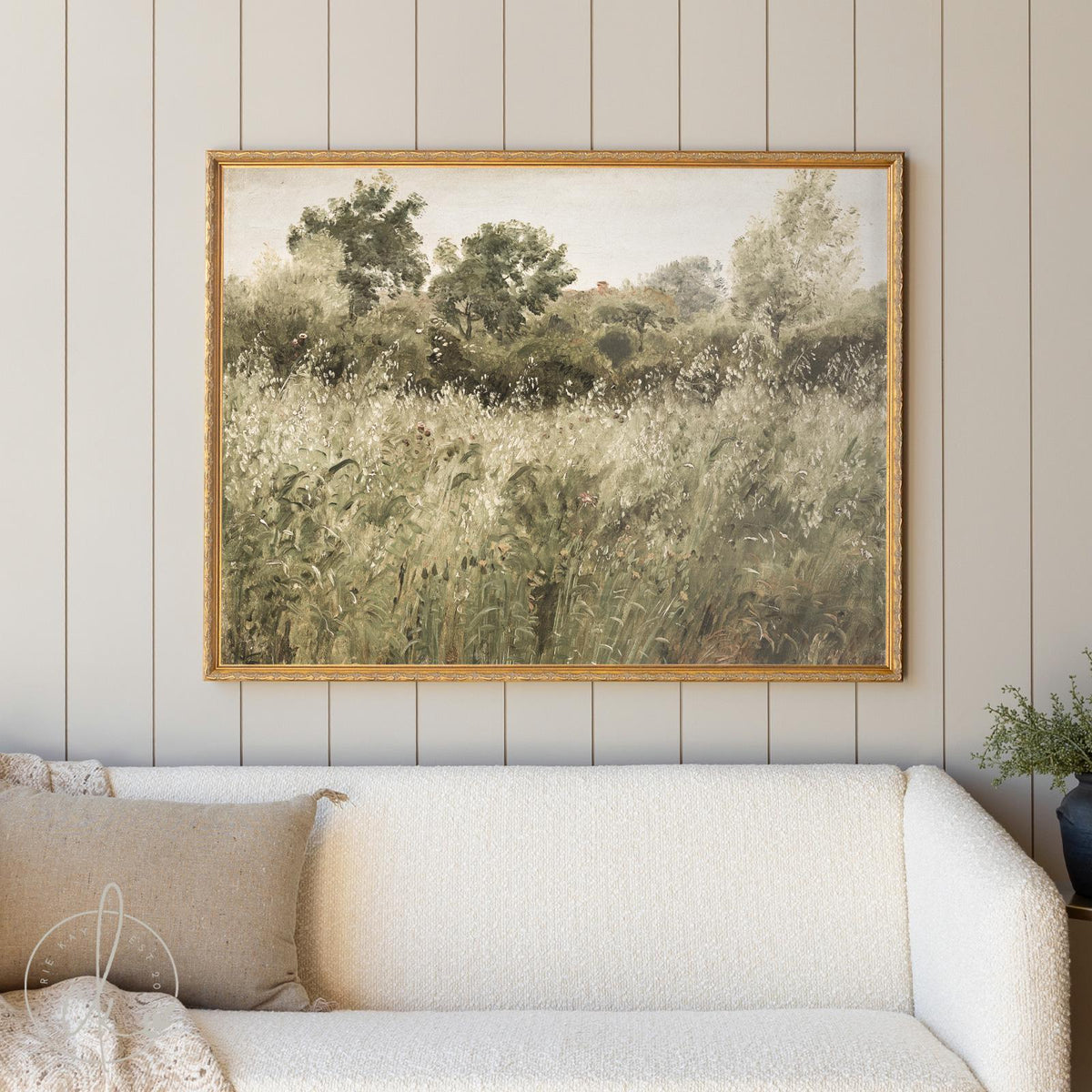 Vintage framed landscape artwork depicting a serene countryside scene with tall grasses, wildflowers, and lush green trees, set against a soft sky. The painting is displayed above a cream boucle sofa in a cozy, neutral-toned living room with vertical shiplap walls, creating a warm and inviting rustic aesthetic.