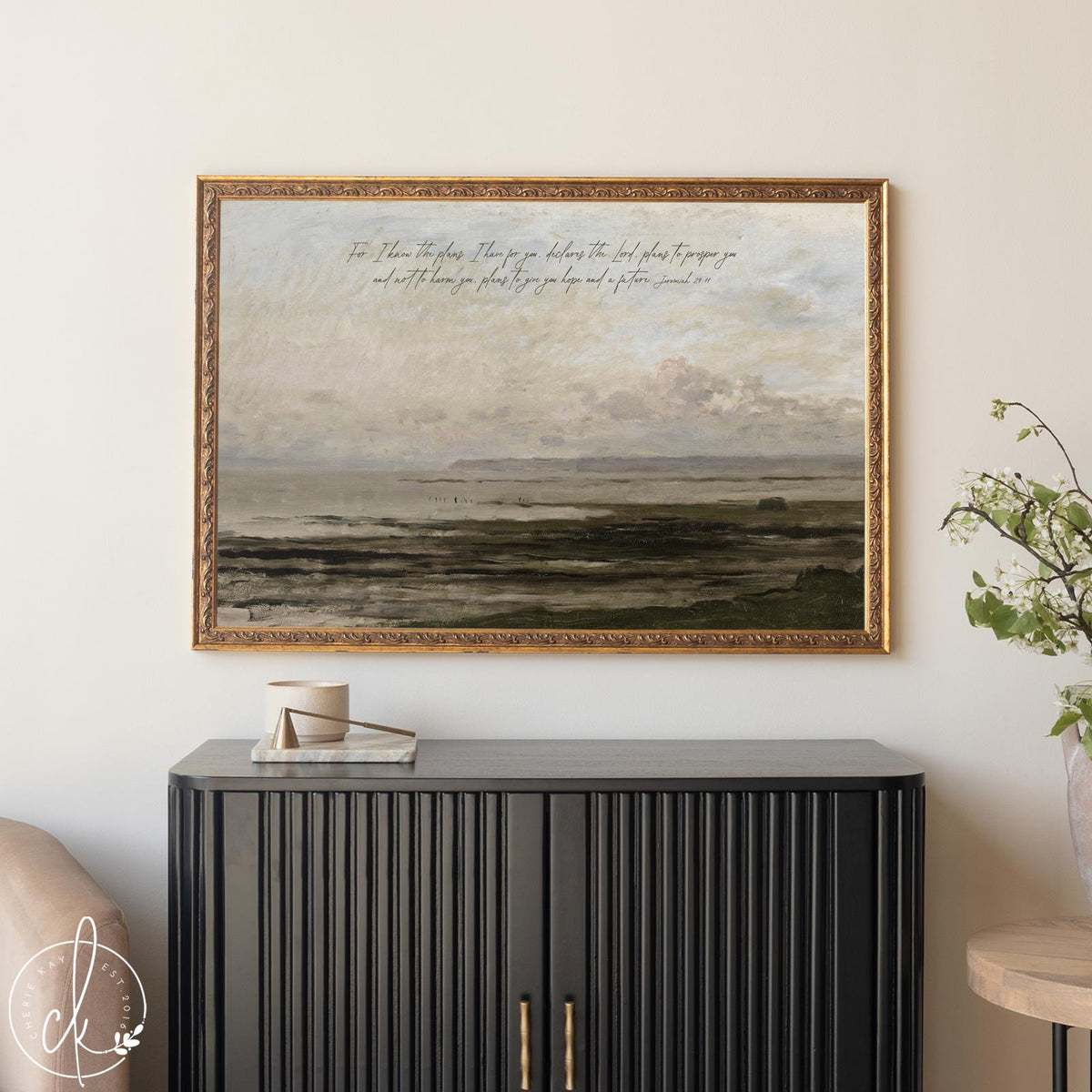 Framed landscape artwork hanging above a modern black cabinet, depicting a serene coastal scene with soft waves, a distant horizon, and pastel sky. Handwritten scripture from Jeremiah 29:11 overlays the top portion of the painting. Decor accents and a leafy branch complete the elegant, minimal room setting.