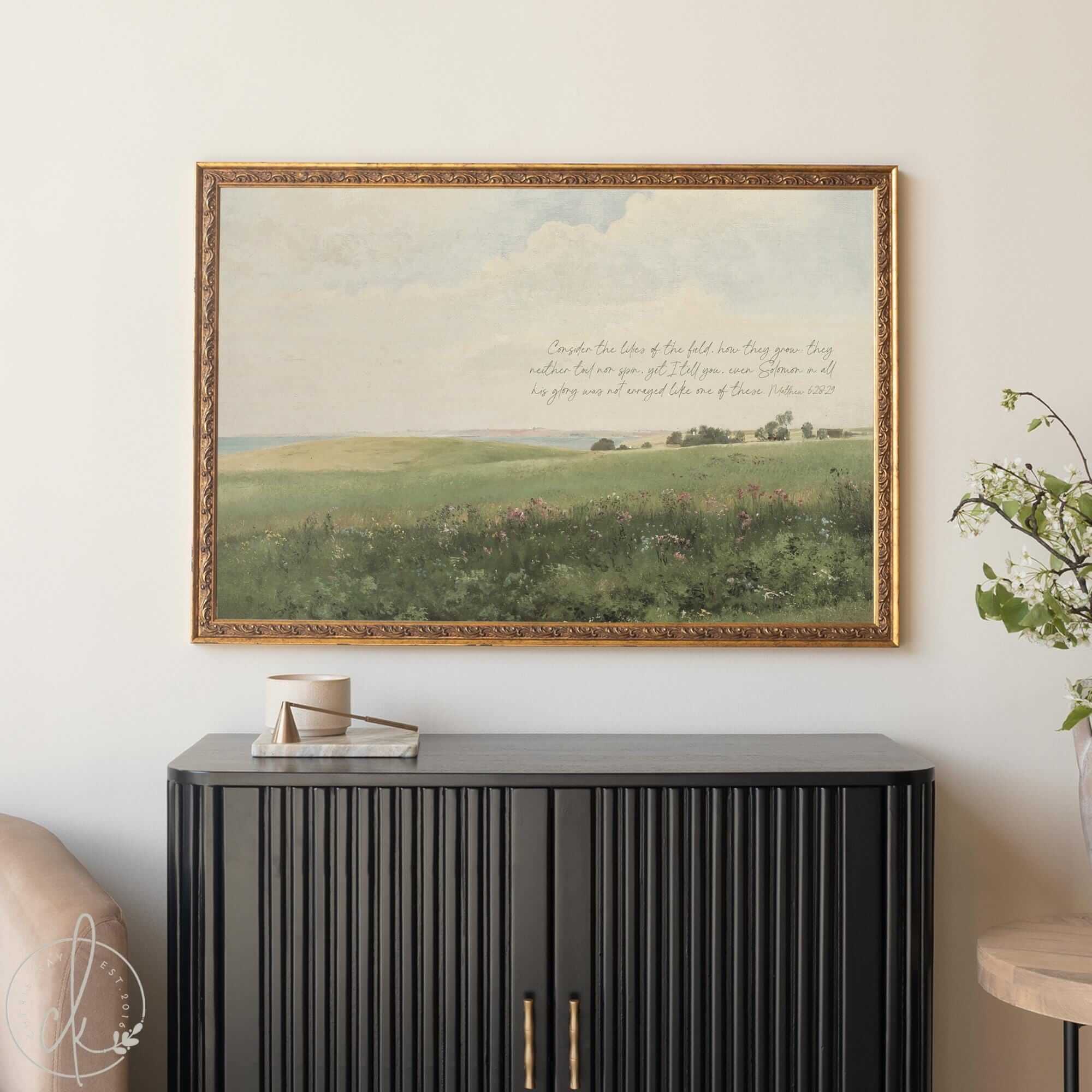 Large wall art with Scripture featuring a soft landscape of a wildflower field with the Bible verse from Matthew 6:28 in elegant script, styled above a black sideboard in a cozy home setting.