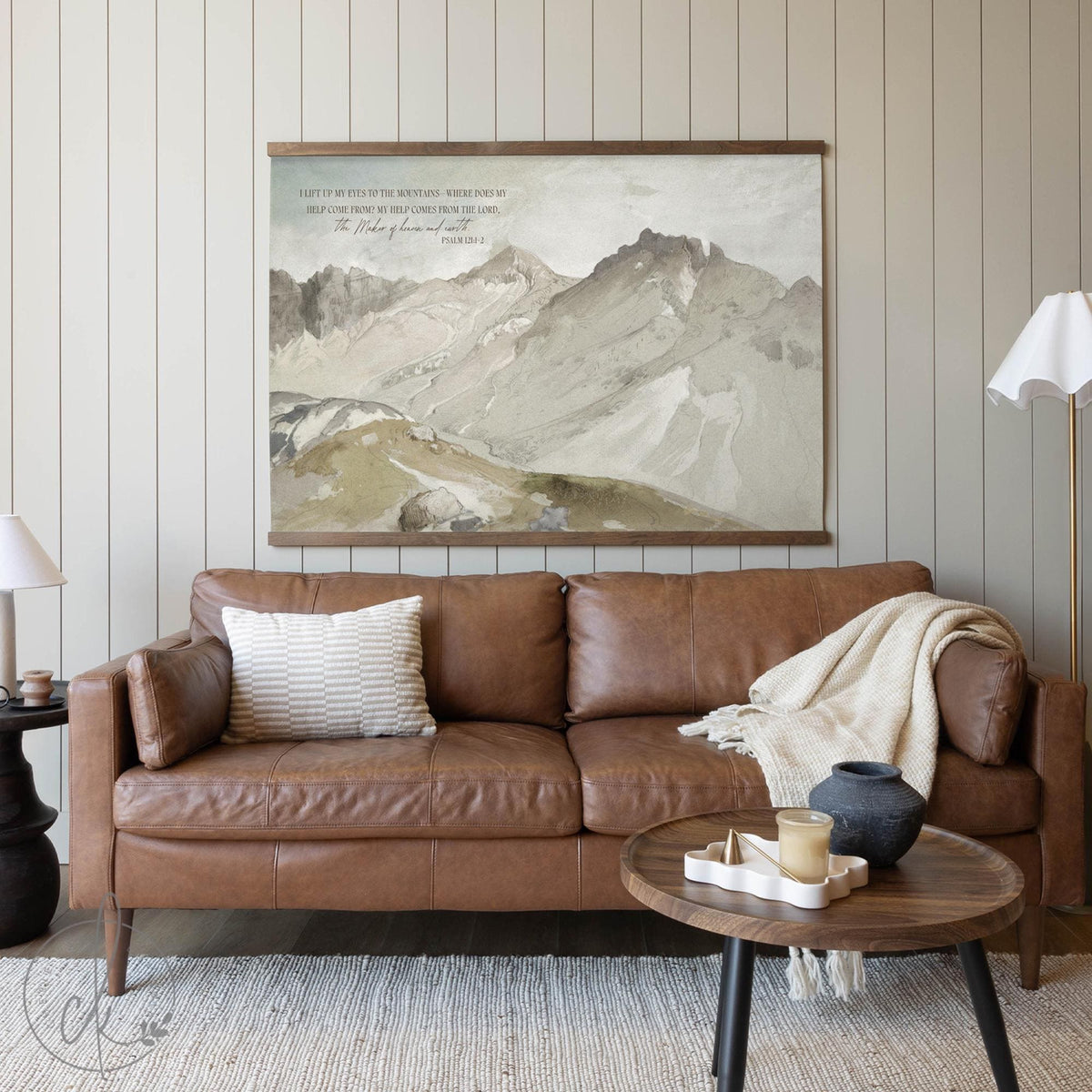 Framed Christian mountain wall art print with Psalm 121 scripture, displayed above a leather sofa in a cozy living room setting.