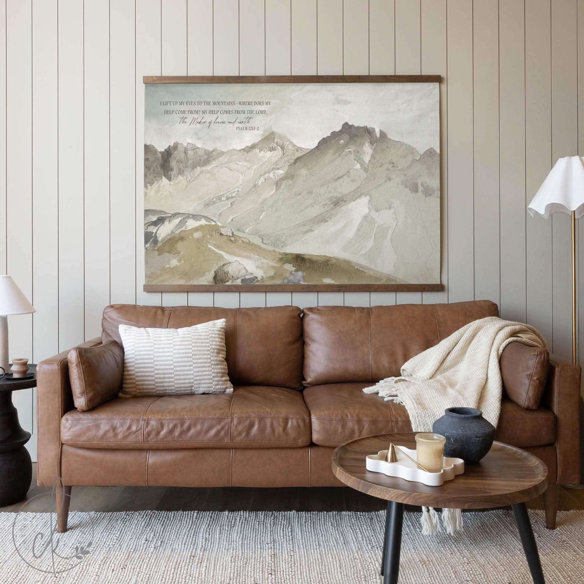 Framed Christian mountain wall art print with Psalm 121 scripture, displayed above a leather sofa in a cozy living room setting.