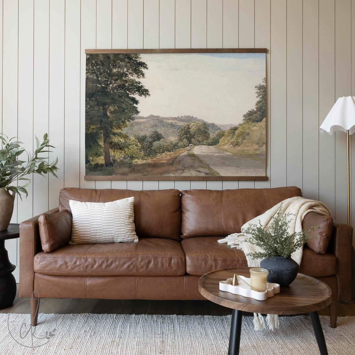 A cozy living room with a brown leather sofa, a textured pillow, and a blanket draped over the arm. A round wooden coffee table with simple decor sits in front, and a wall-mounted art piece of a scenic road and trees hangs above.