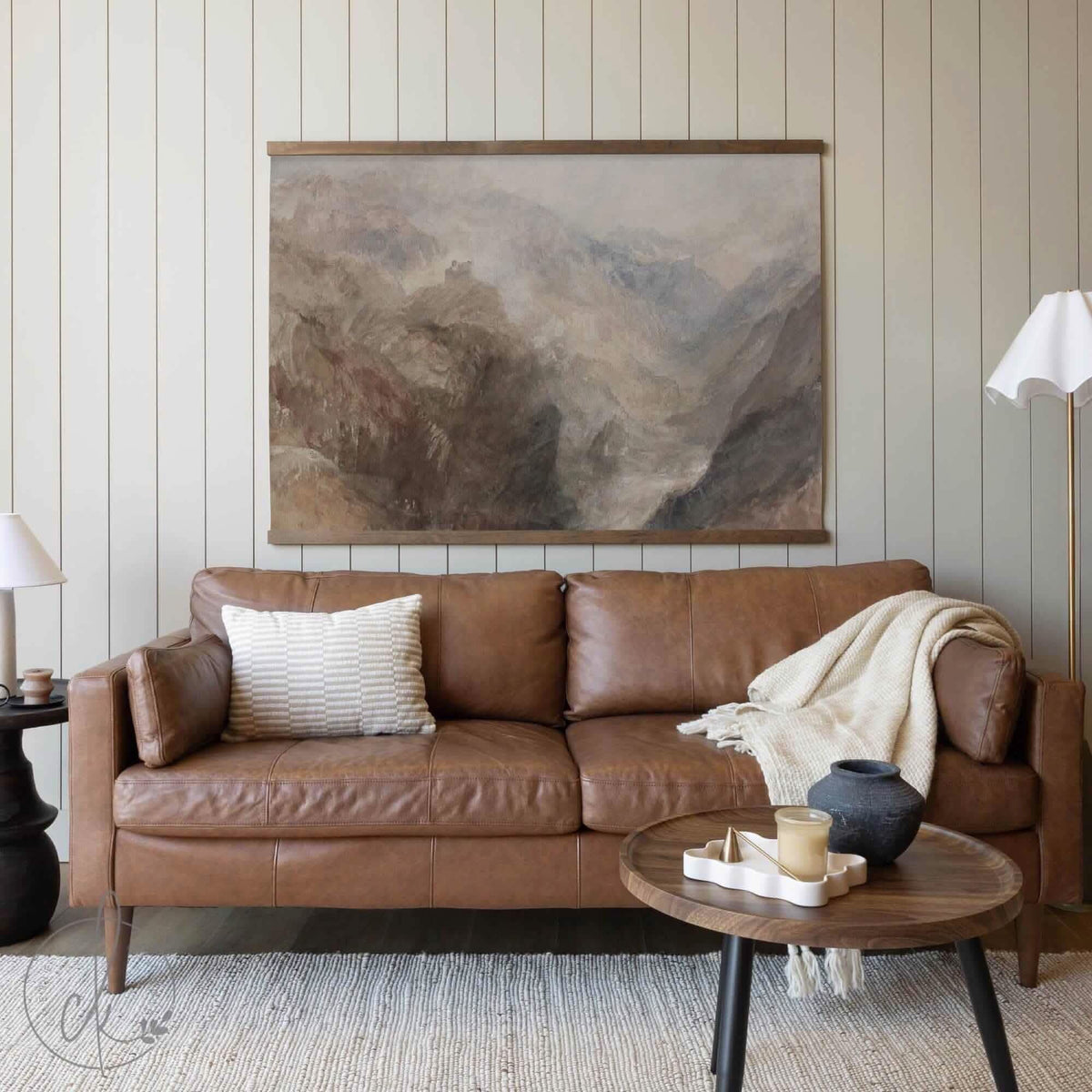 A cozy living room displaying a large mountain landscape canvas tapestry in neutral tones above a brown leather sofa, accented by a light-colored throw blanket and pillow.