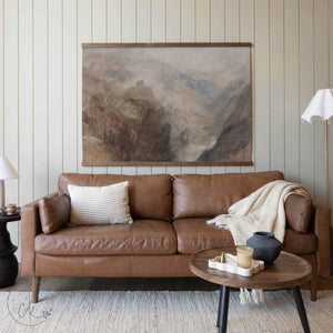 A cozy living room displaying a large mountain landscape canvas tapestry in neutral tones above a brown leather sofa, accented by a light-colored throw blanket and pillow.