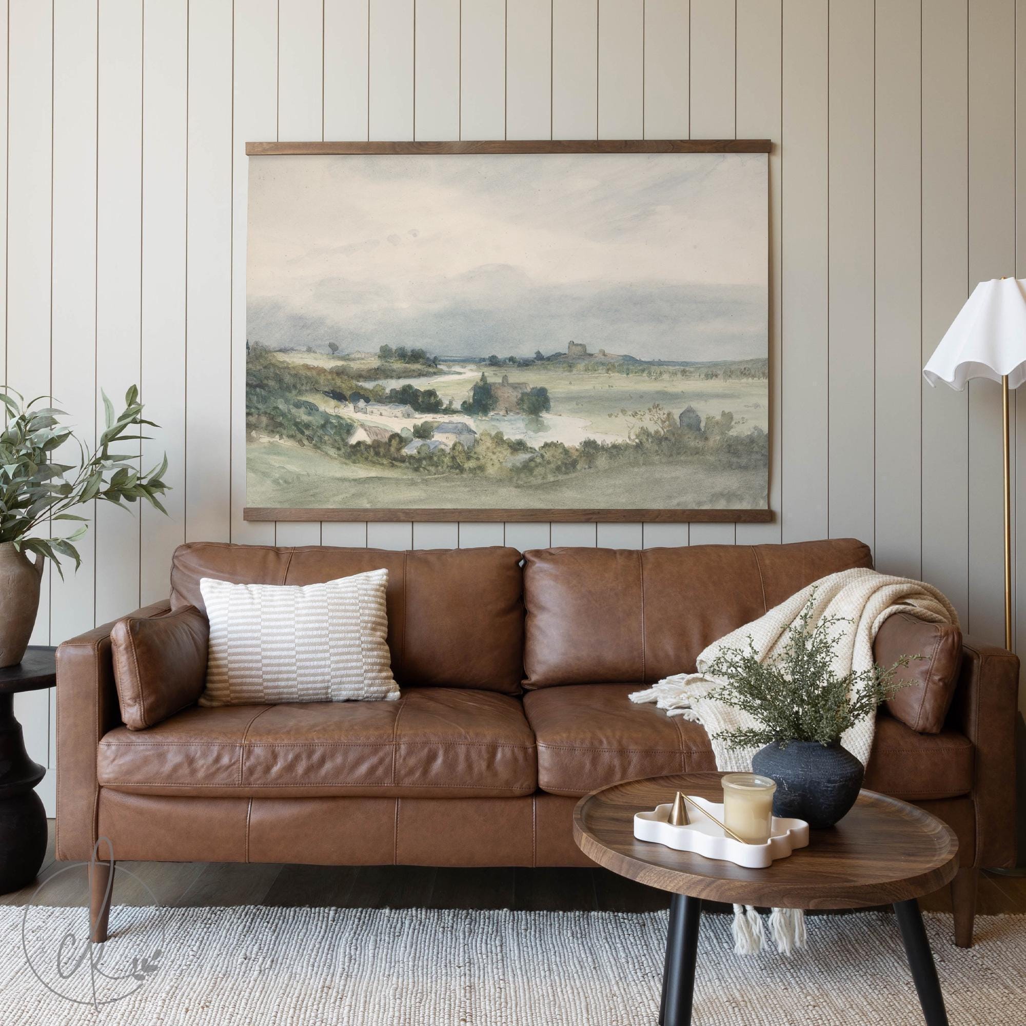 A cozy living room with a brown leather sofa, a wooden coffee table, and a potted plant, featuring a large landscape painting on the wall.