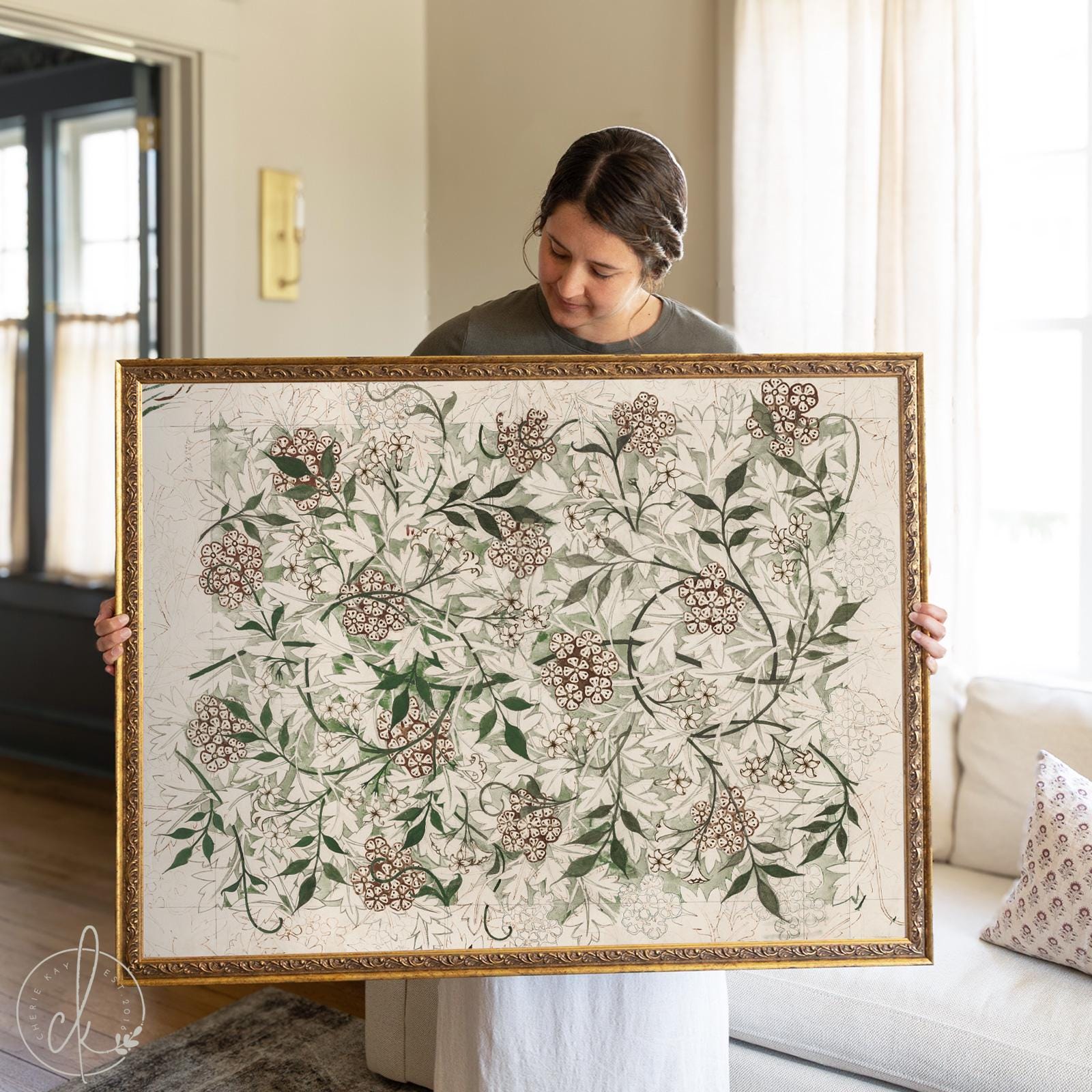 Woman holding a large framed vintage botanical textile artwork with intricate green leaves and floral patterns, displayed in a softly lit living room.