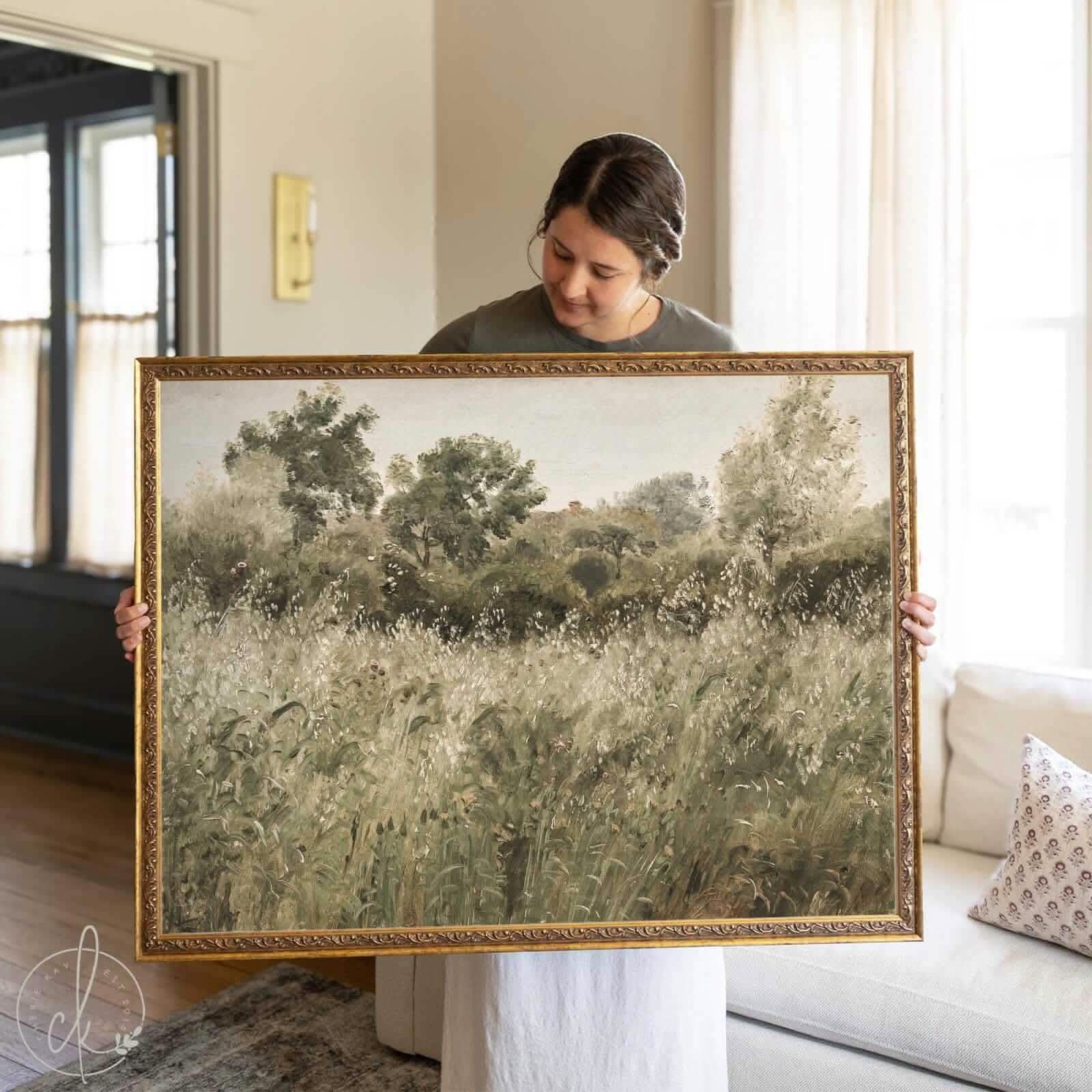Woman holding a large framed landscape painting featuring a lush green meadow with tall grasses and wildflowers, set against a backdrop of dense trees under a pale sky. The artwork is in an ornate gold frame, and the scene is captured in a cozy, naturally lit living room.