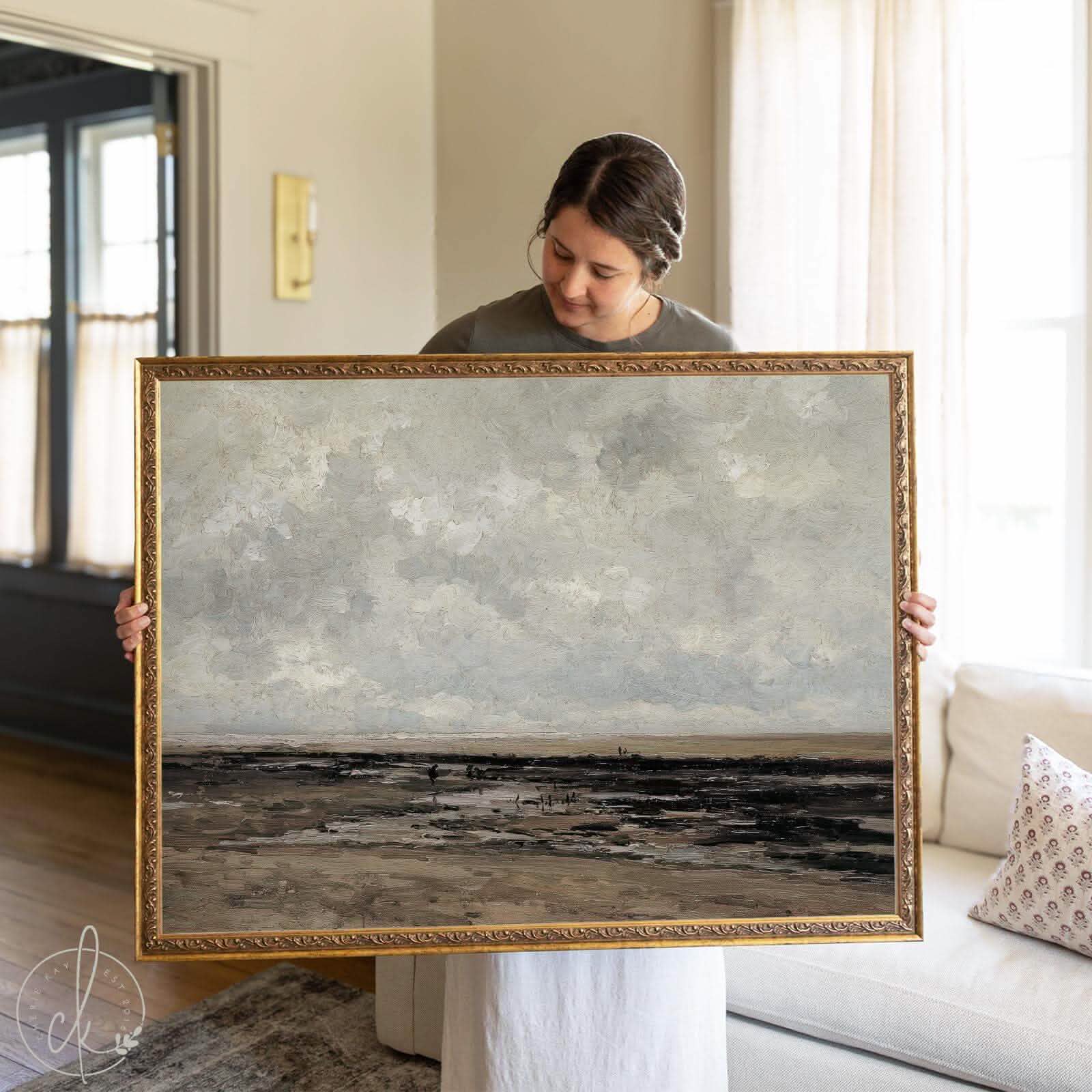 Woman holding a large vintage framed landscape painting featuring a moody coastal scene in earthy tones, ideal wall art for traditional or modern vintage home decor.
