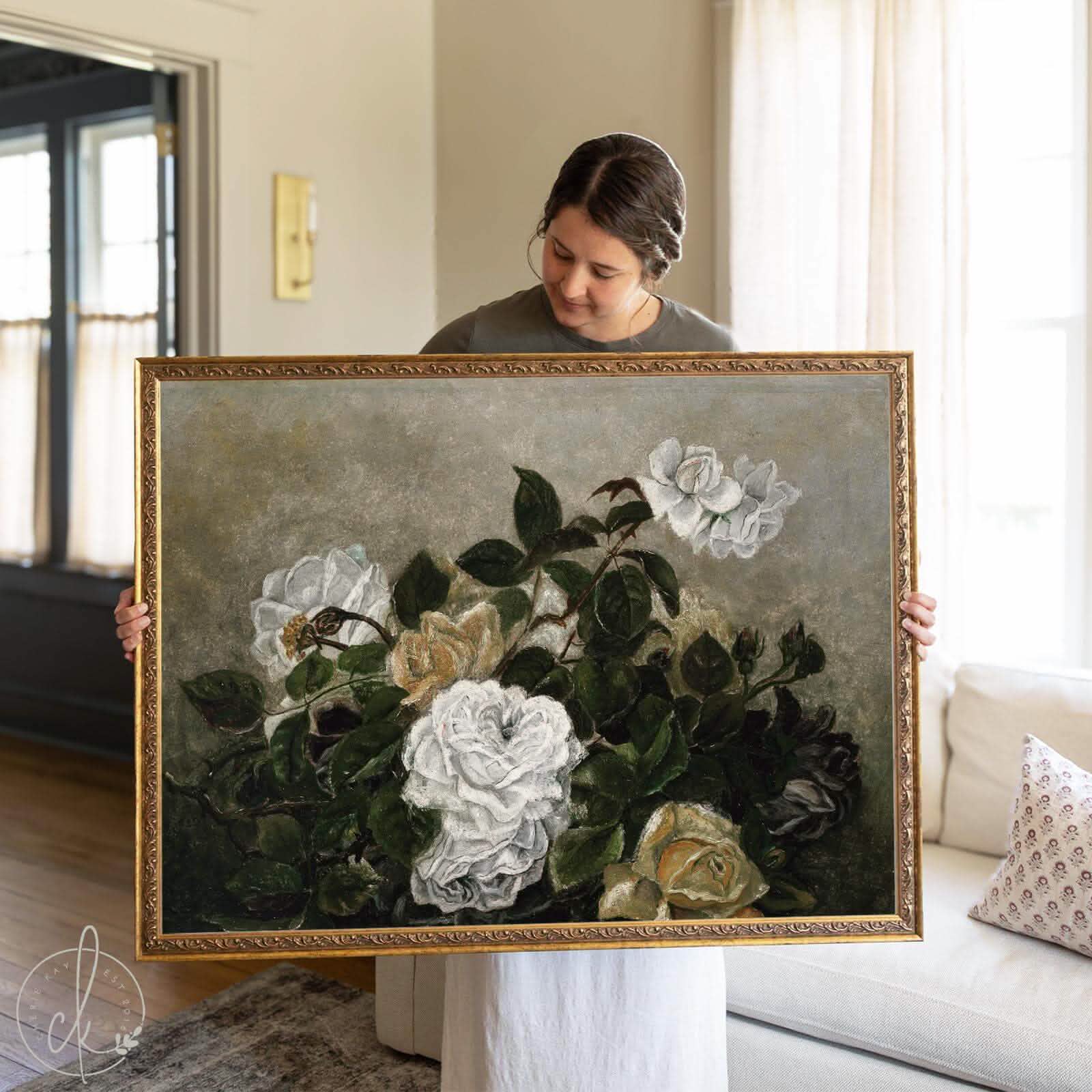 Woman holding a large framed vintage botanical art print featuring white and yellow roses with deep green leaves, set against a muted gray background. The floral painting is housed in an ornate gold frame and styled in a cozy, softly lit living room.