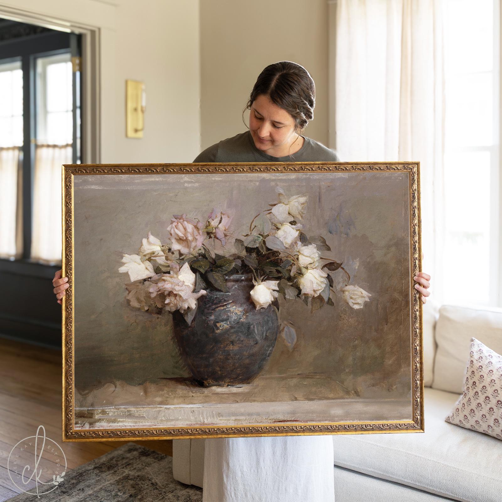 A woman holds a large framed vintage botanical art print featuring soft white and blush roses in a rustic blue ceramic vase. The floral still life has a muted, antique finish and is ideal for farmhouse or traditional home decor styles.