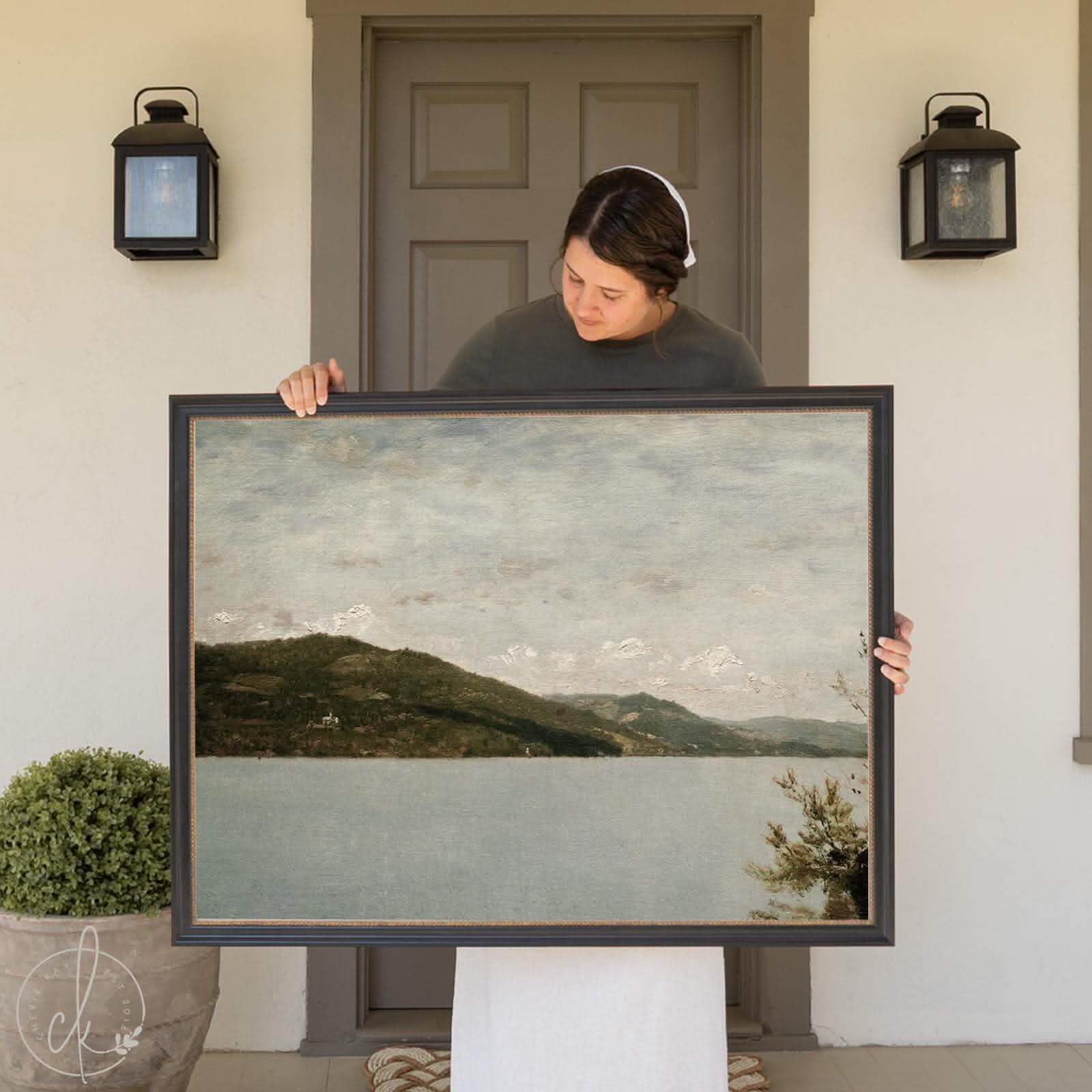A woman stands in front of a house, holding a large vintage framed oil painting of a lake and mountain. The artwork features a calm, muted blue lake bordered by a tree-covered hillside, with a range of distant mountains fading into the soft, cloudy sky. The frame has a classic, dark finish that complements the natural tones of the artwork.