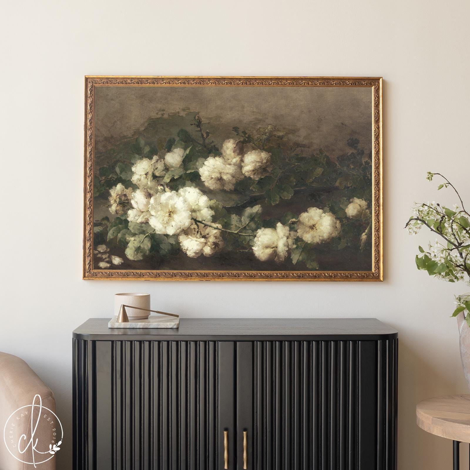 Framed vintage floral painting of white roses with green foliage, hanging above a modern black cabinet in a neutral-toned living room. The artwork features an ornate gold frame and a muted, antique-style background, complementing the minimal, elegant decor.