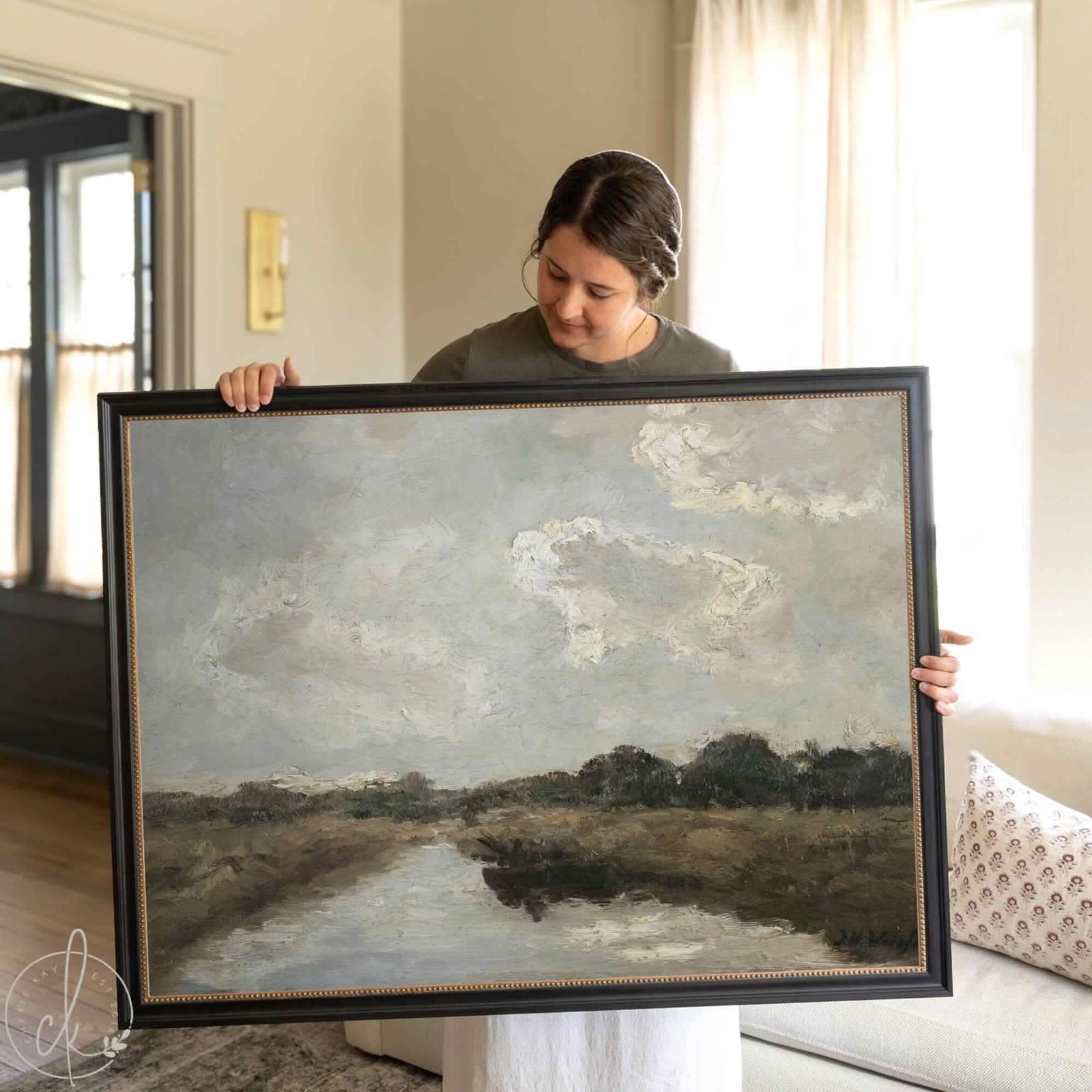 Woman holding a large vintage framed art print featuring a cloudy landscape with neutral tones, rustic scenery, and textured brushstrokes, perfect for traditional or farmhouse home decor.
