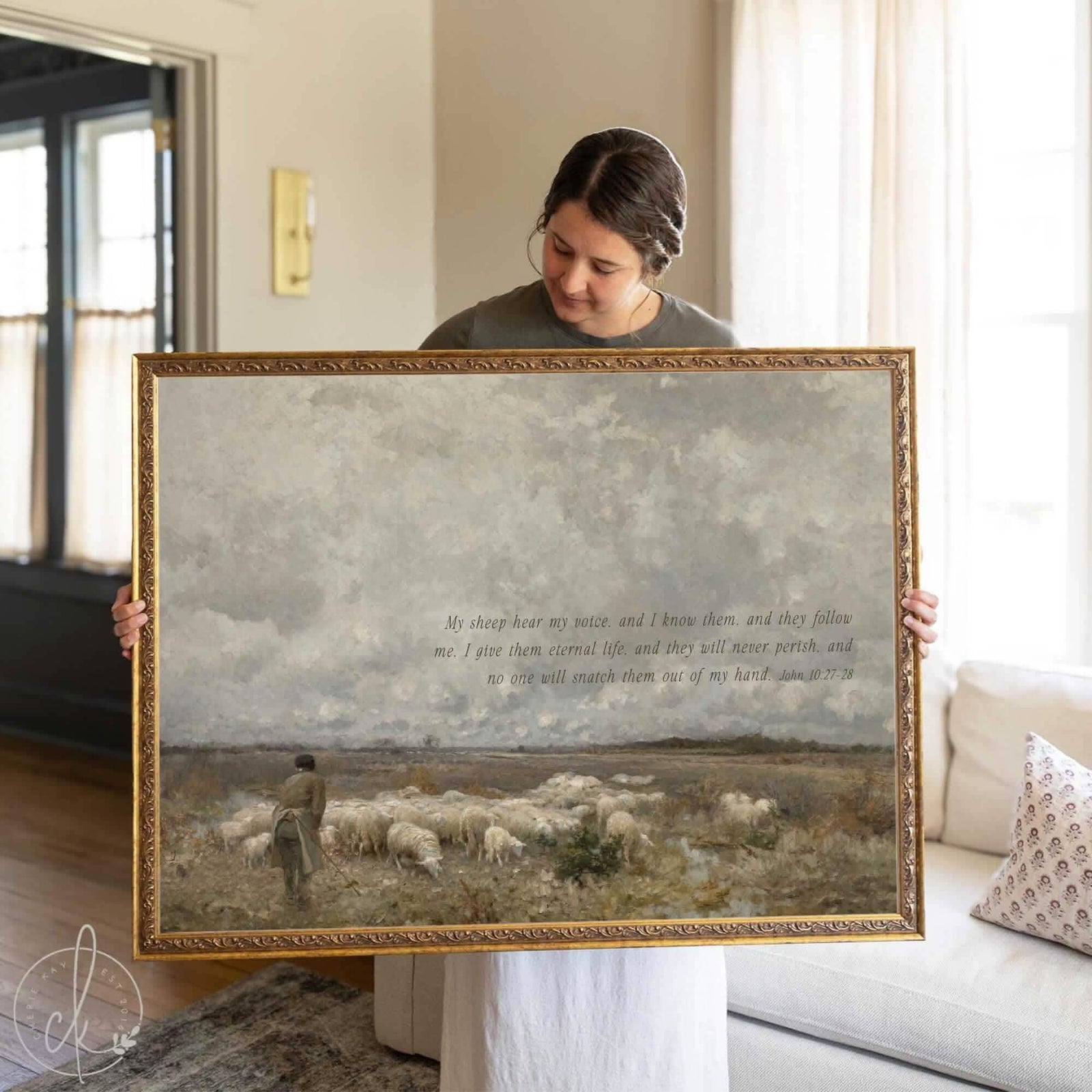 Woman holding a large framed Christian wall art featuring a shepherd with sheep under a cloudy sky and the Bible verse John 10:27–28; vintage-inspired wall art for faith-based home decor.