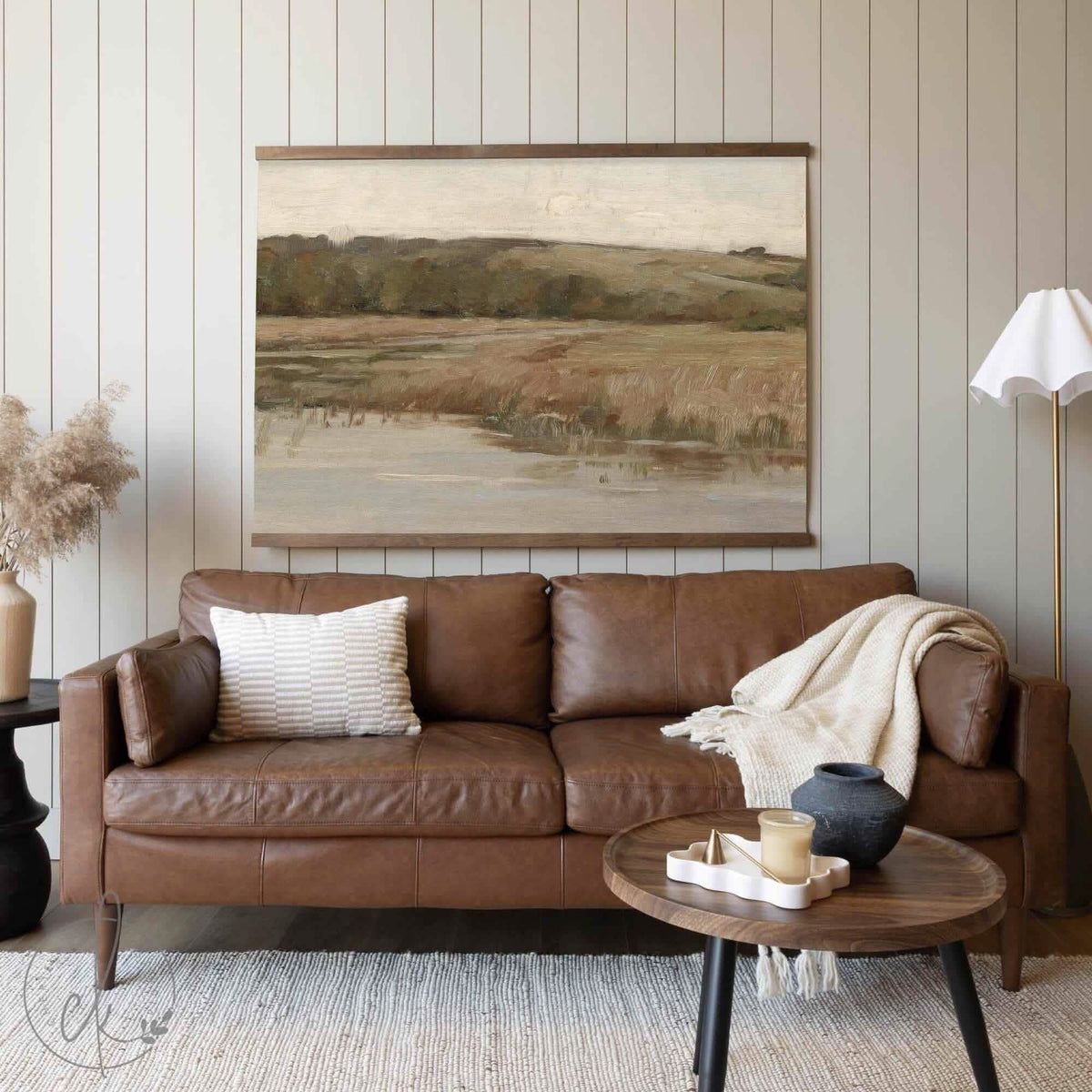 A large, neutral-toned landscape tapestry featuring rolling hills and grassy fields, hung above a warm brown leather sofa in a cozy living room setting.