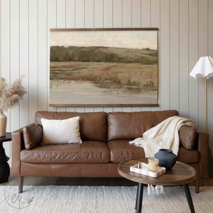 A large, neutral-toned landscape tapestry featuring rolling hills and grassy fields, hung above a warm brown leather sofa in a cozy living room setting.