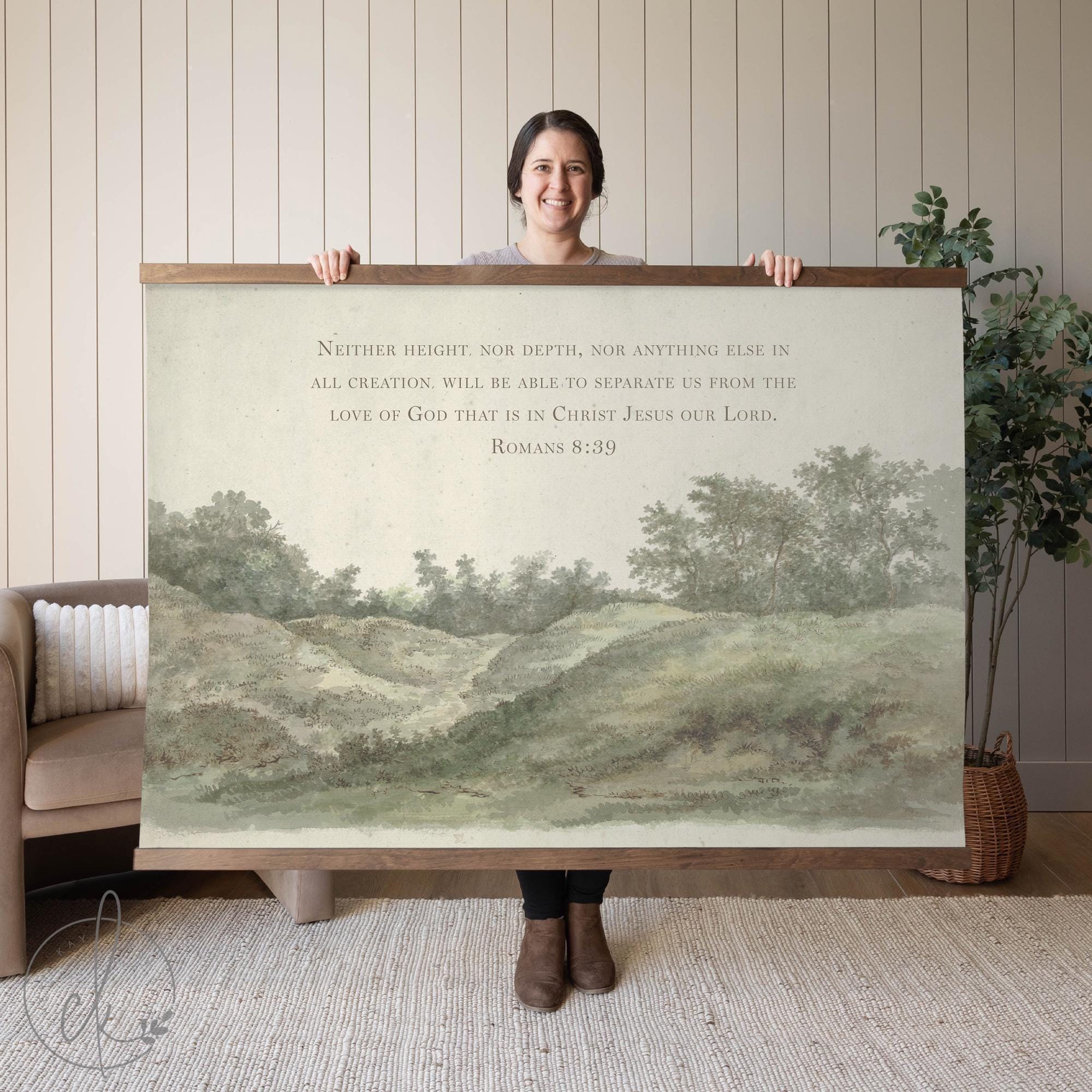 A person holding wall art of a countryside landscape with a verse from Romans 8:39. The background features a sofa and a potted plant.
