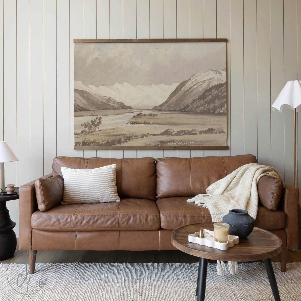 A large neutral-tone mountain landscape tapestry hanging above a caramel-brown leather sofa, accented by a cream pillow, cozy throw blanket, and a simple round coffee table in a softly lit living room setting.