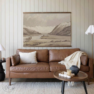 A large neutral-tone mountain landscape tapestry hanging above a caramel-brown leather sofa, accented by a cream pillow, cozy throw blanket, and a simple round coffee table in a softly lit living room setting.