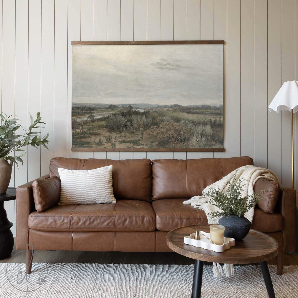 Staged living room with a brown leather sofa, neutral décor accents, and a large tapestry displaying a serene countryside landscape in muted earth tones.