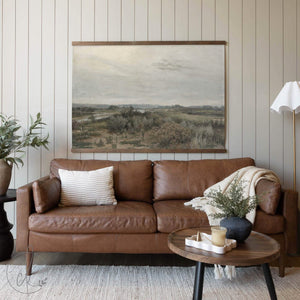 Staged living room with a brown leather sofa, neutral décor accents, and a large tapestry displaying a serene countryside landscape in muted earth tones.