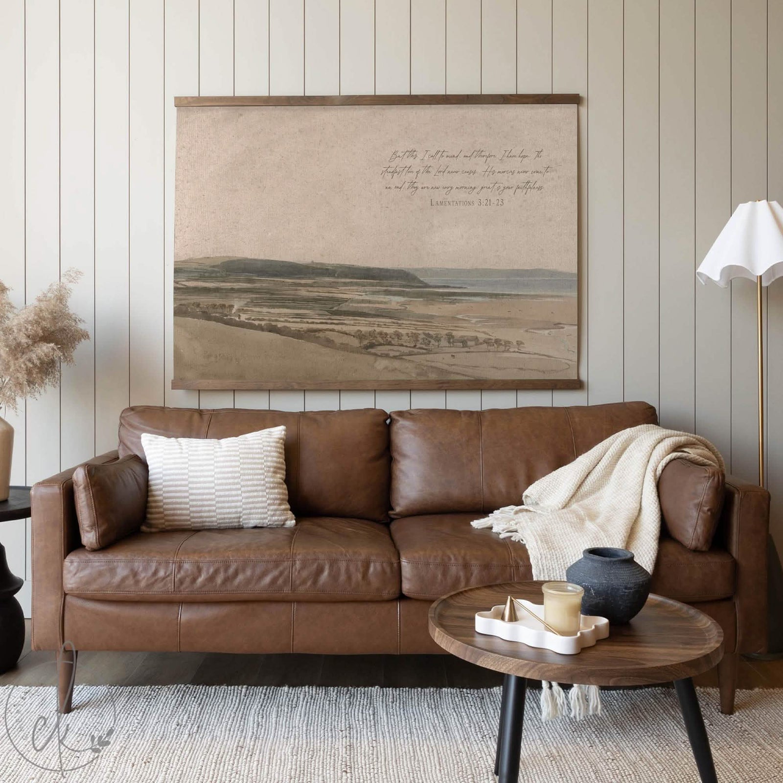 Large watercolor landscape tapestry with vintage coastal scenery and Lamentations scripture, styled above a brown leather sofa in a cozy, neutral-toned living room.