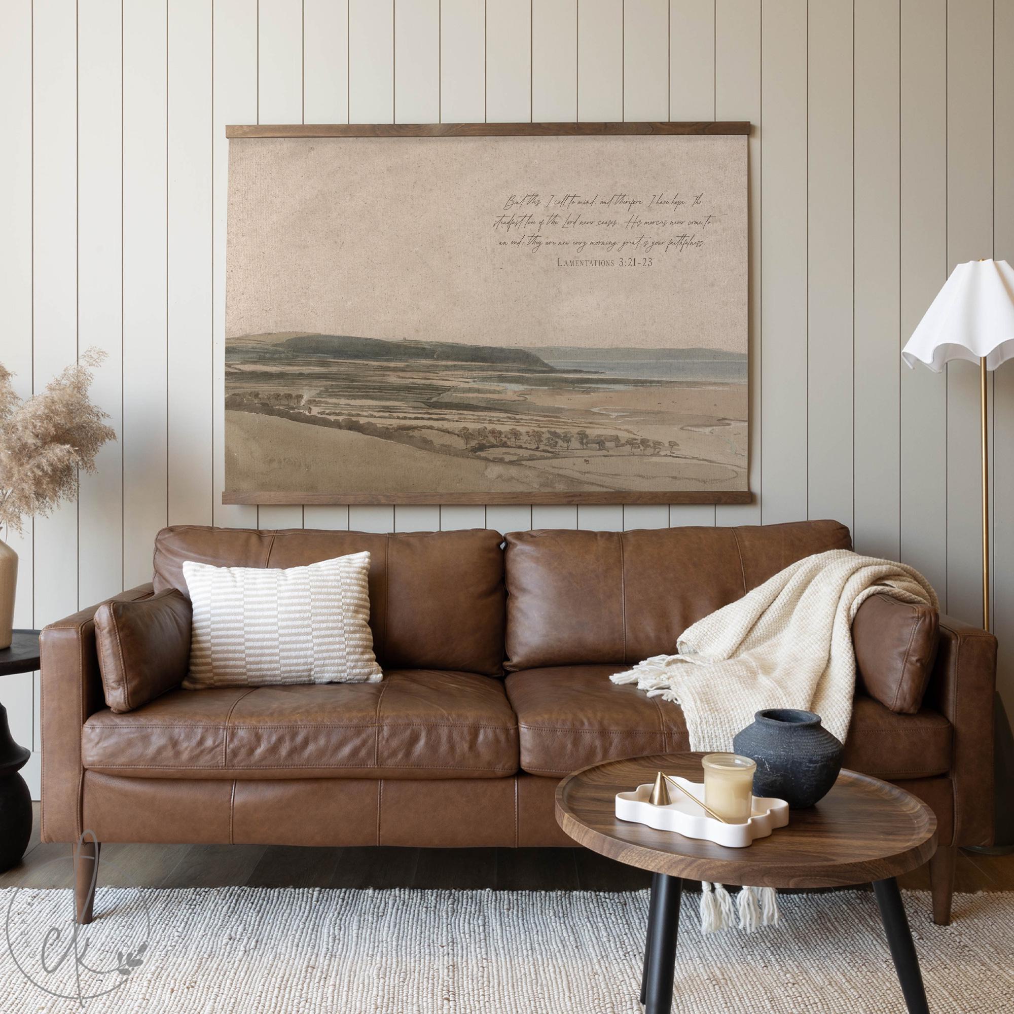Large watercolor landscape tapestry with vintage coastal scenery and Lamentations scripture, styled above a brown leather sofa in a cozy, neutral-toned living room.