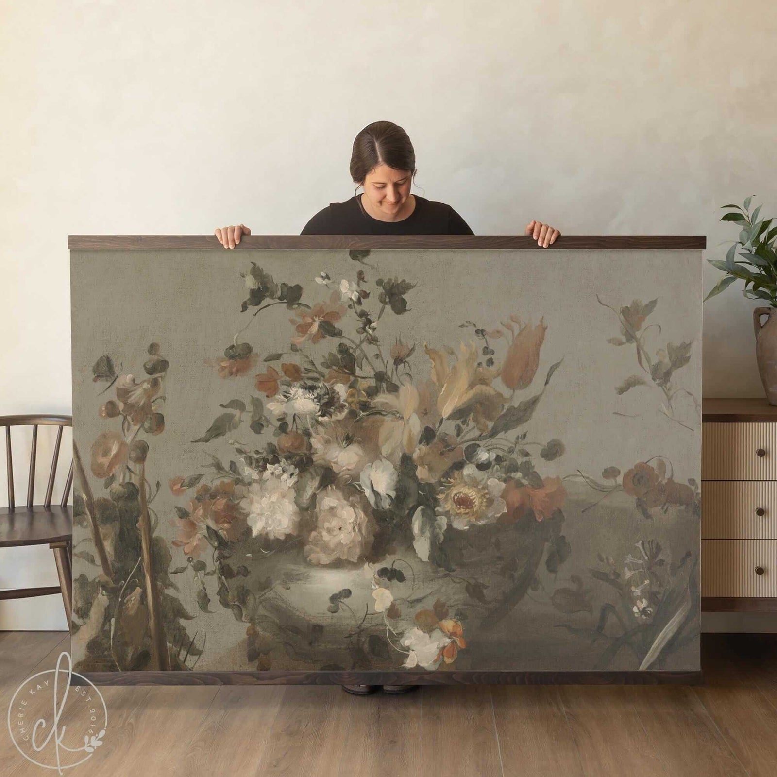 Vintage floral still life canvas wall hanging in muted botanical tones, shown oversized and held by a woman in a modern farmhouse interior.