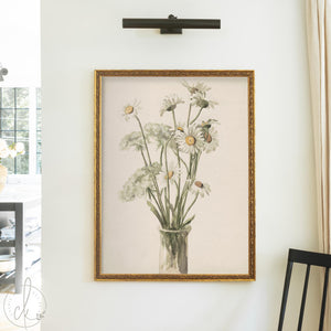 Daisy wall decor with white daisies and Queen Anne's lace in a glass vase in an ornate gold wood frame on a cream wall.