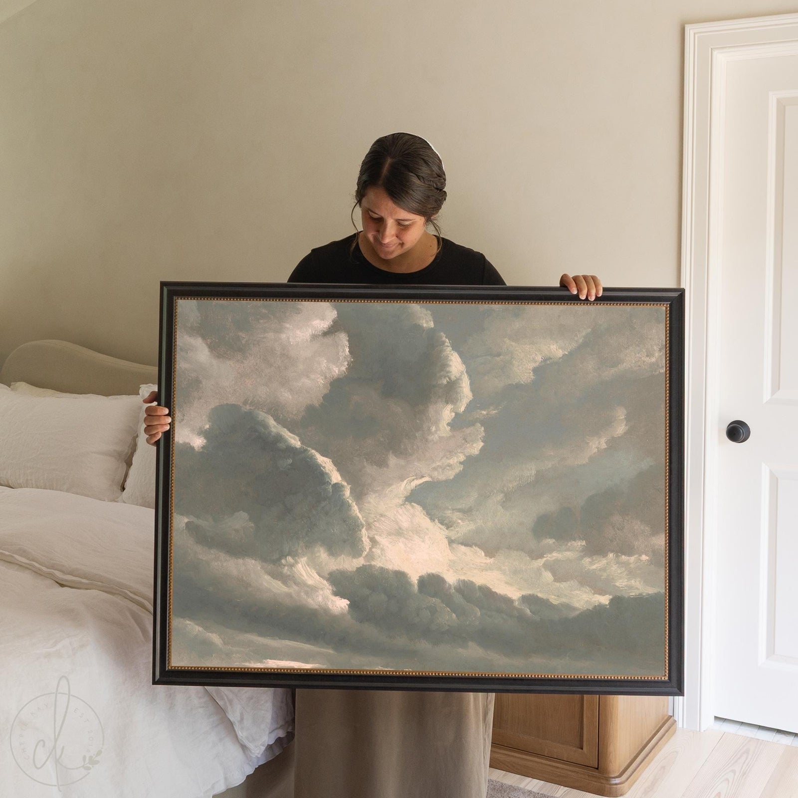 Vintage sky painting of sunlit clouds in a black frame with gold bead trim, displayed above a bed in a neutral bedroom.