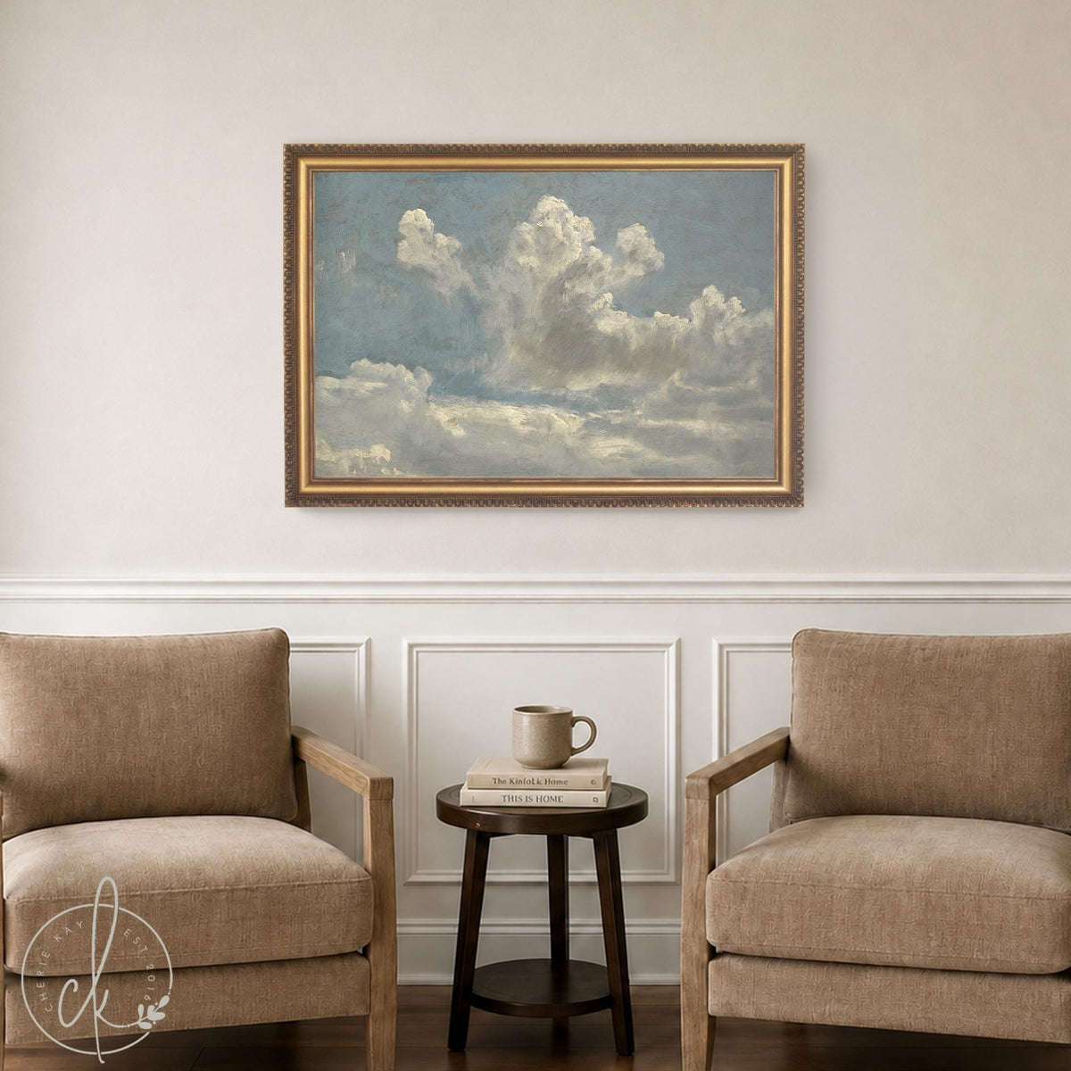Cloud art print in an ornate gold frame, hung above a side table between two armchairs in a neutral living room