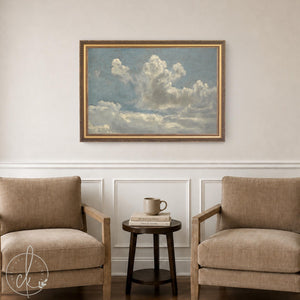 Cloud art print in an ornate gold frame, hung above a side table between two armchairs in a neutral living room
