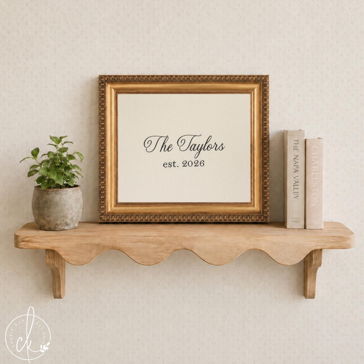 Embroidered wall art family name sign, black script name and est. year on cream fabric in an ornate gold frame on a shelf
