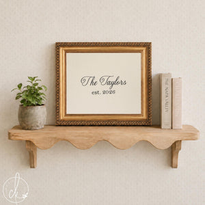 Embroidered wall art family name sign, black script name and est. year on cream fabric in an ornate gold frame on a shelf