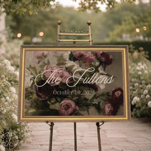 Wedding ceremony sign with last name and date over a vintage rose painting, ornate gold frame, shown on an easel in a garden.
