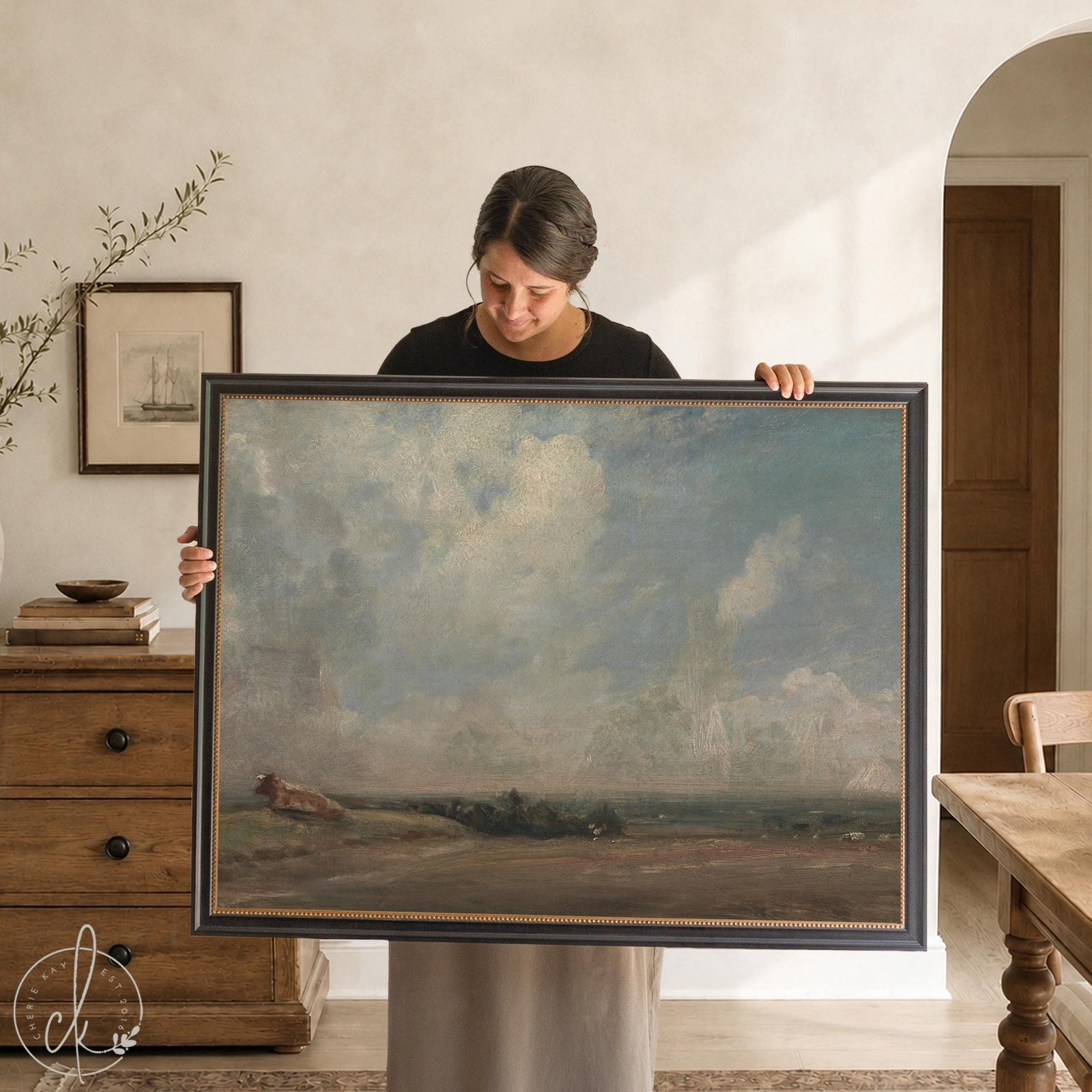 Woman holds a large pastoral painting print of a cloudy sky over an open field in a black and gold frame