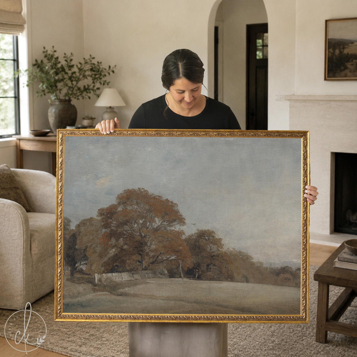 Woman holding vintage fall art on framed canvas, amber trees by a stone wall in an ornate gold frame.